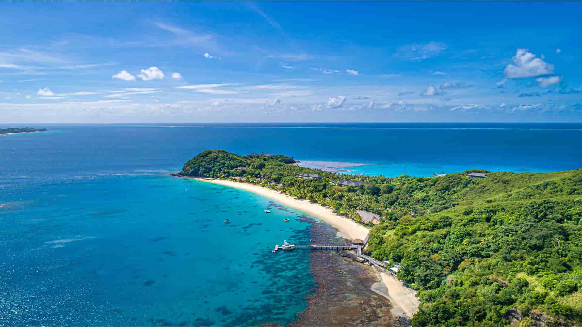 An aerial view of Kadavu Island in Fiji, showcasing a tropical coastline with a pristine white-sand beach, a pier extending over clear turquoise water, and lush green forest leading up to the ocean.