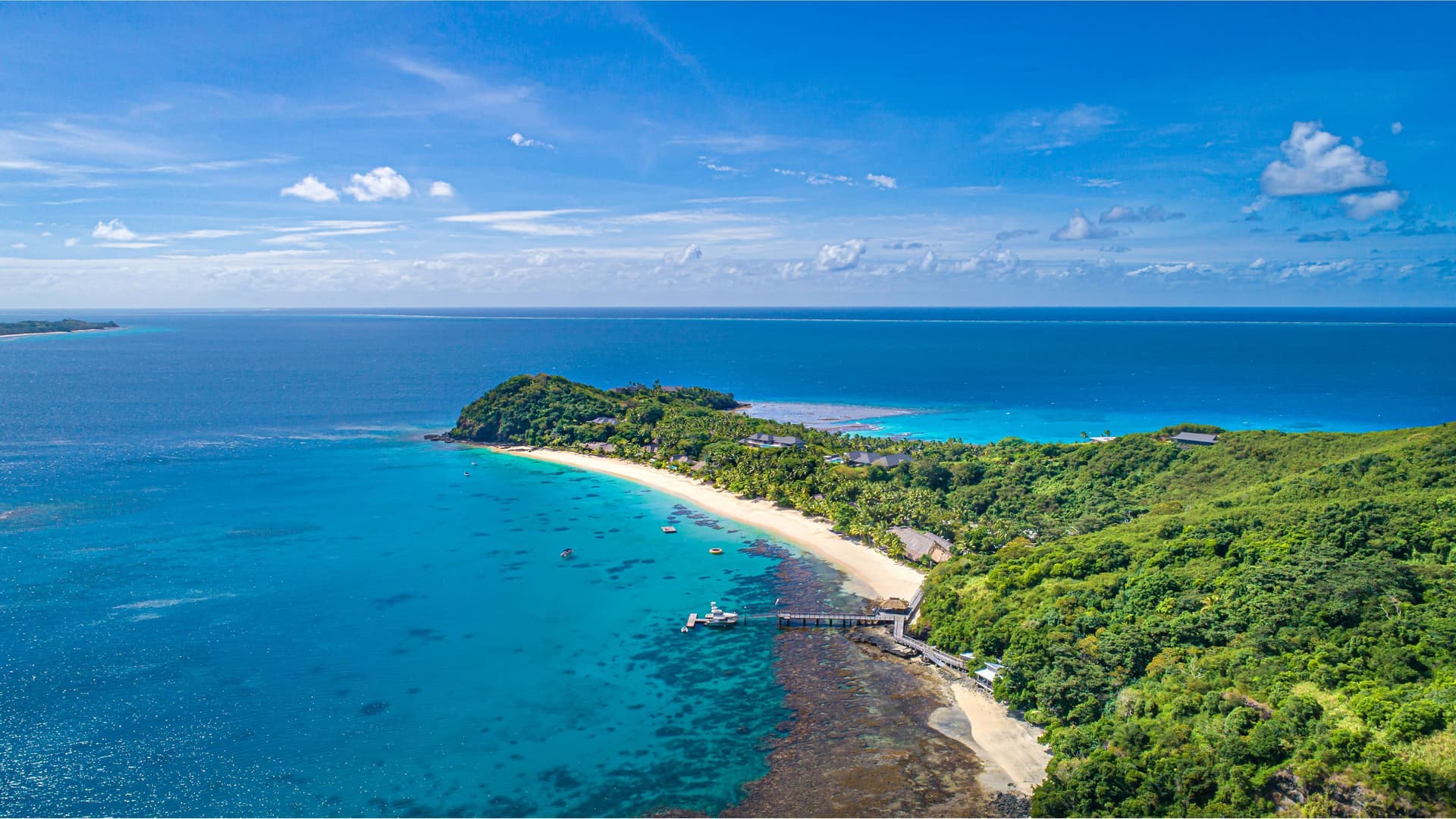 An aerial view of Kadavu Island in Fiji, showcasing a tropical coastline with a pristine white-sand beach, a pier extending over clear turquoise water, and lush green forest leading up to the ocean.