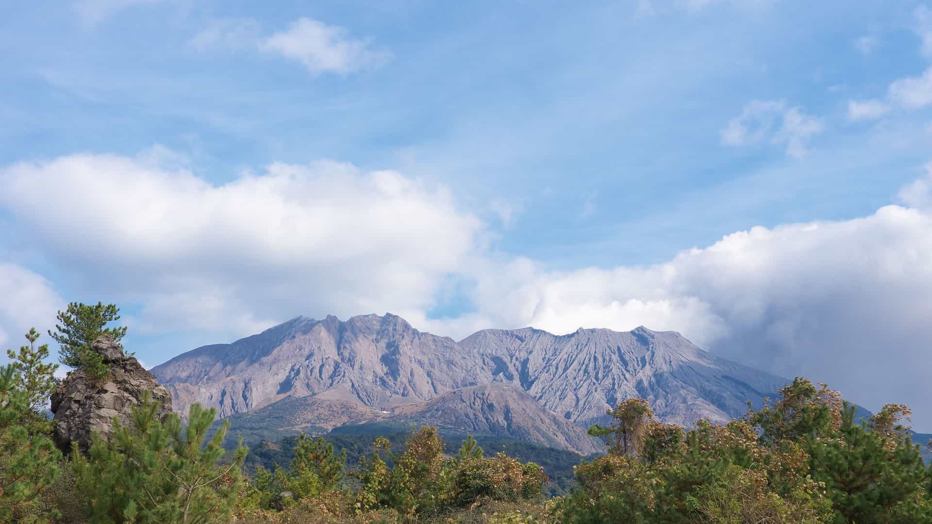 A striking view of the active Sakurajima volcano and its peaks, rising behind a foreground of green trees, in the bay of Kagoshima, Japan.