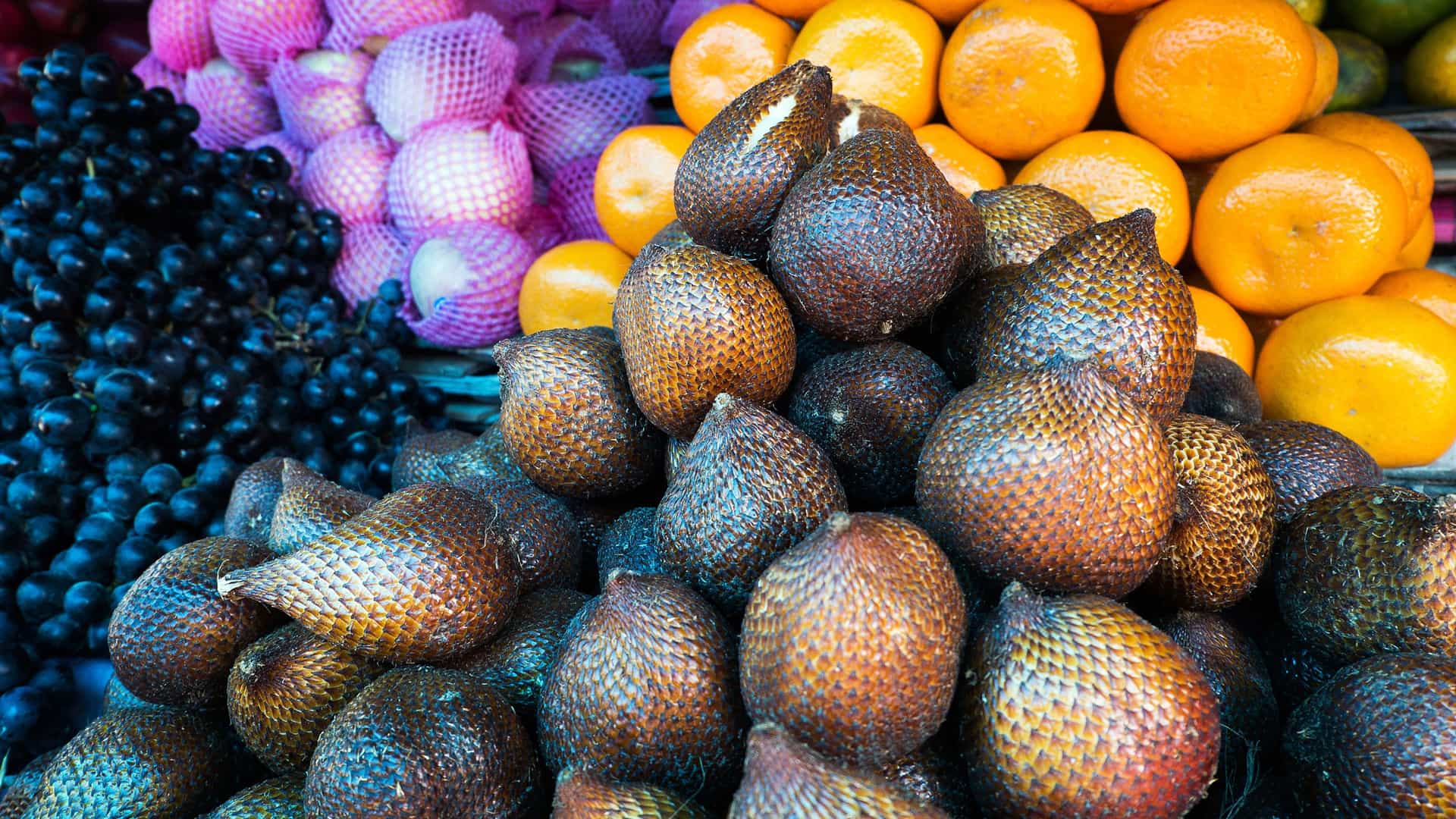 A vibrant close-up of a fruit stall in Kalabahi, Indonesia, showcasing a large pile of Salak (snake fruit) with its unique scaly skin, surrounded by other fruits like grapes and oranges.