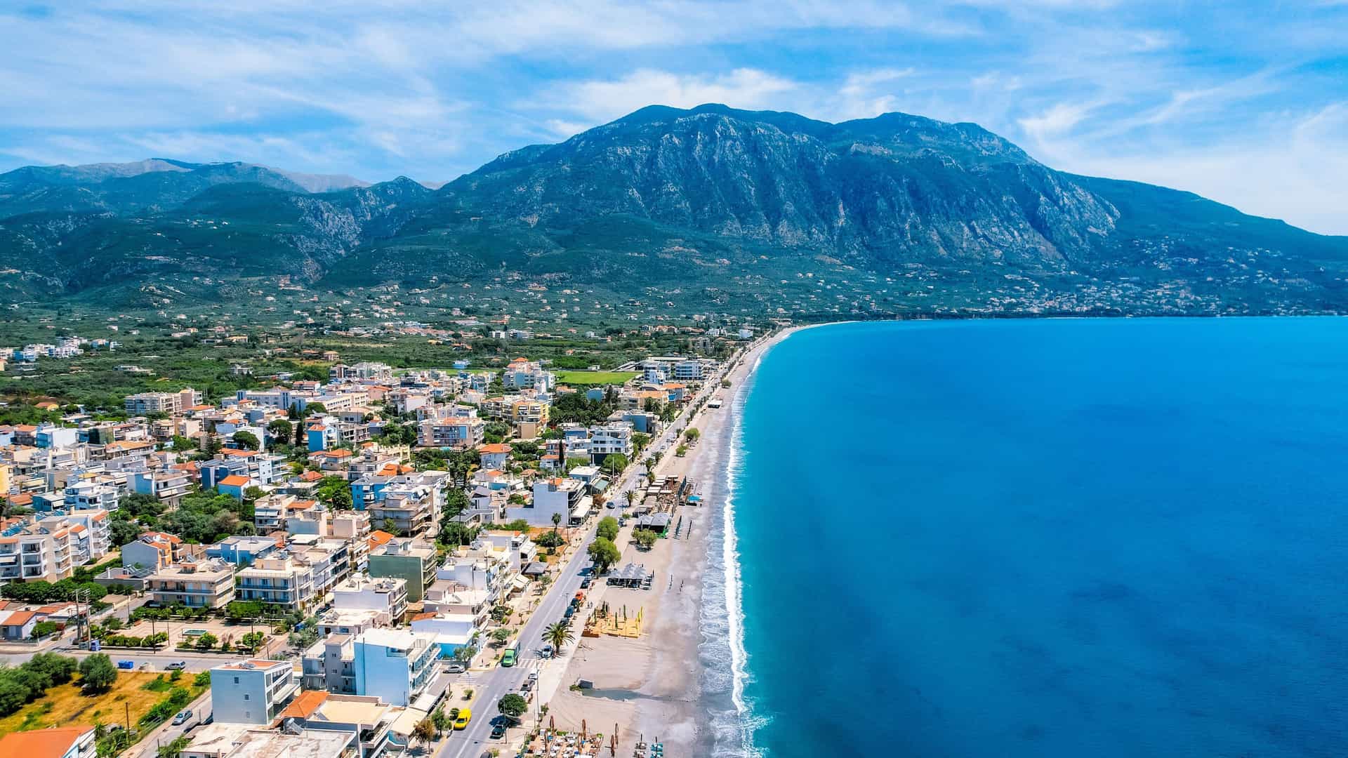 An aerial view of Kalamata, Greece, showing the coastal city and its long sandy beach alongside the deep blue waters of the Messinian Gulf, with Mount Taygetus in the background under a blue sky.