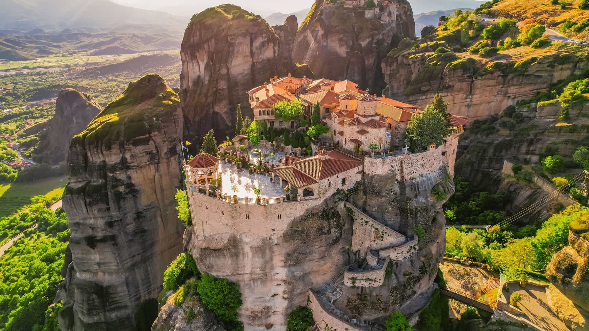 An aerial view of the historic Rousanou Monastery, a UNESCO World Heritage site, perched atop a massive rock formation in Meteora, near Kalabaka, Greece, with surrounding monasteries visible in the distance.