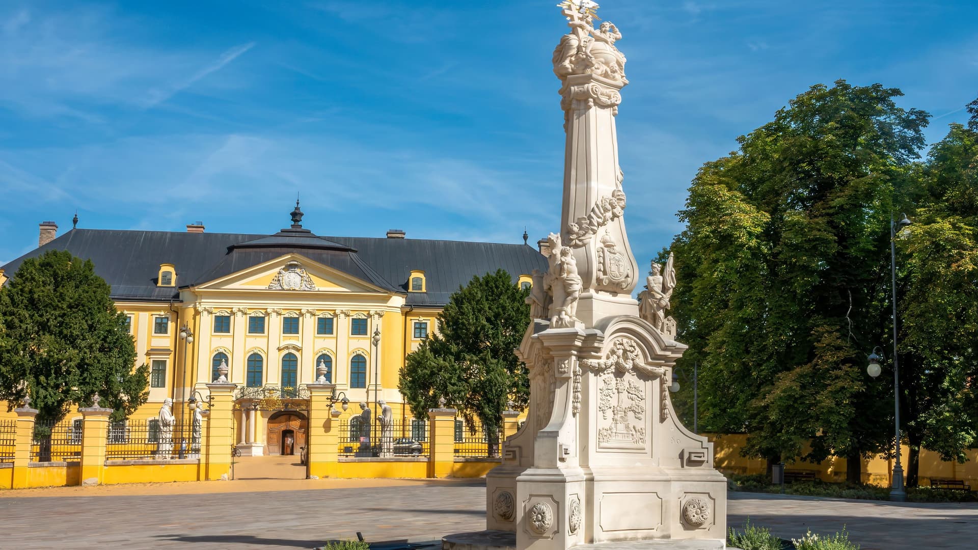 A scenic view of the ornate Holy Trinity Column in the central square of Kalocsa, Hungary, with the historic yellow Archbishop's Palace and lush green trees in the background under a blue sky.