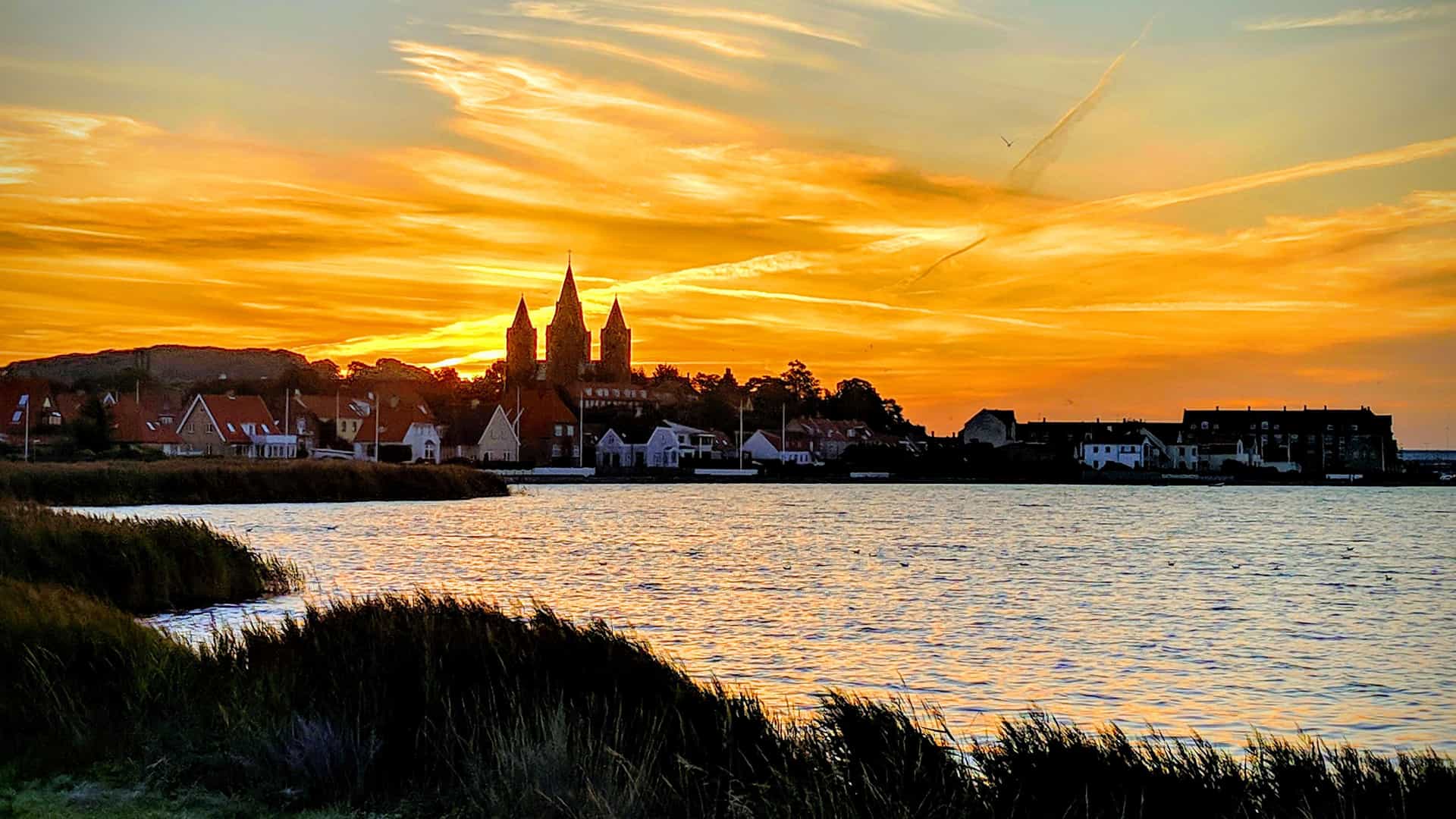 A vibrant sunset over the waterfront town of Kalundborg, Denmark, with the distinctive five-spired Church of Our Lady silhouetted against a brilliant orange and yellow sky, and the bay reflecting the colors.