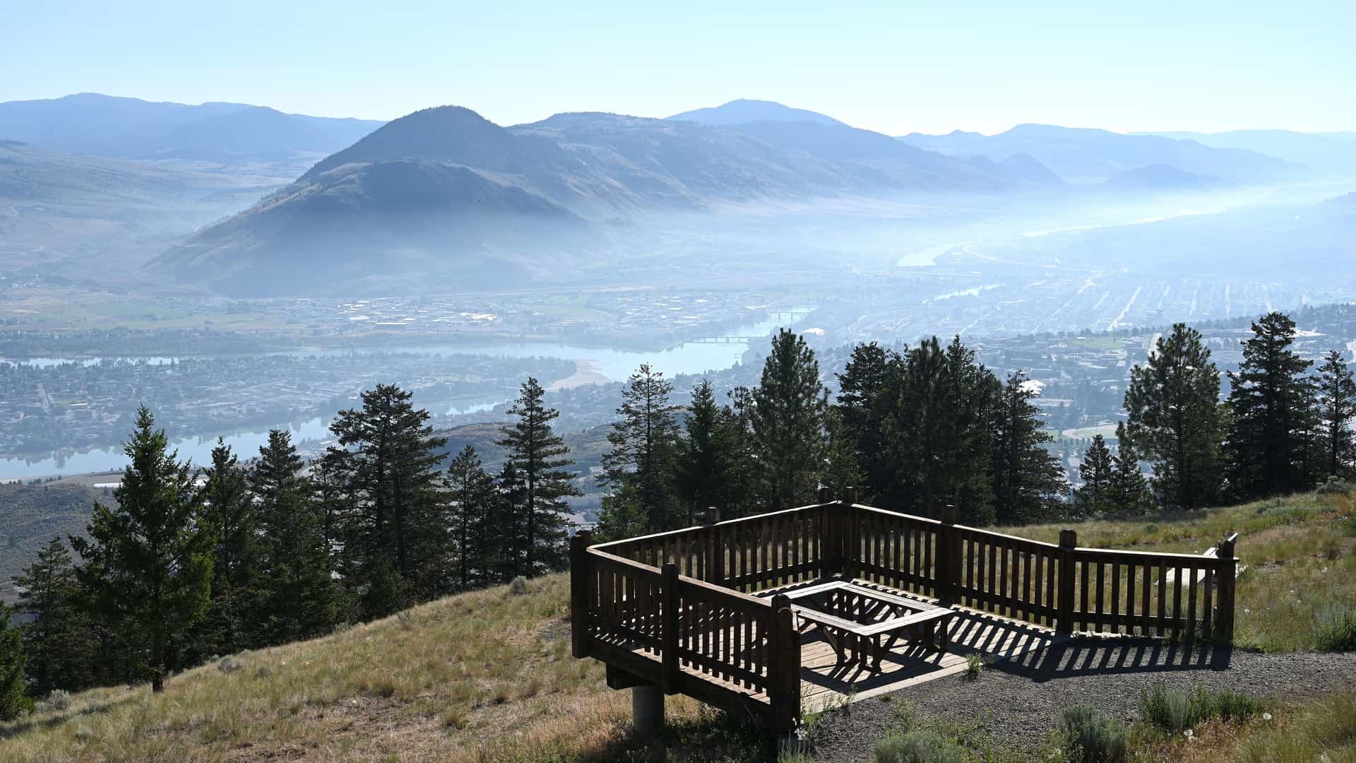 "A beautiful elevated panoramic view of the misty valley of Kamloops, British Columbia, with the North and South Thompson rivers flowing through the city, and a wooden viewing platform in the foreground. "