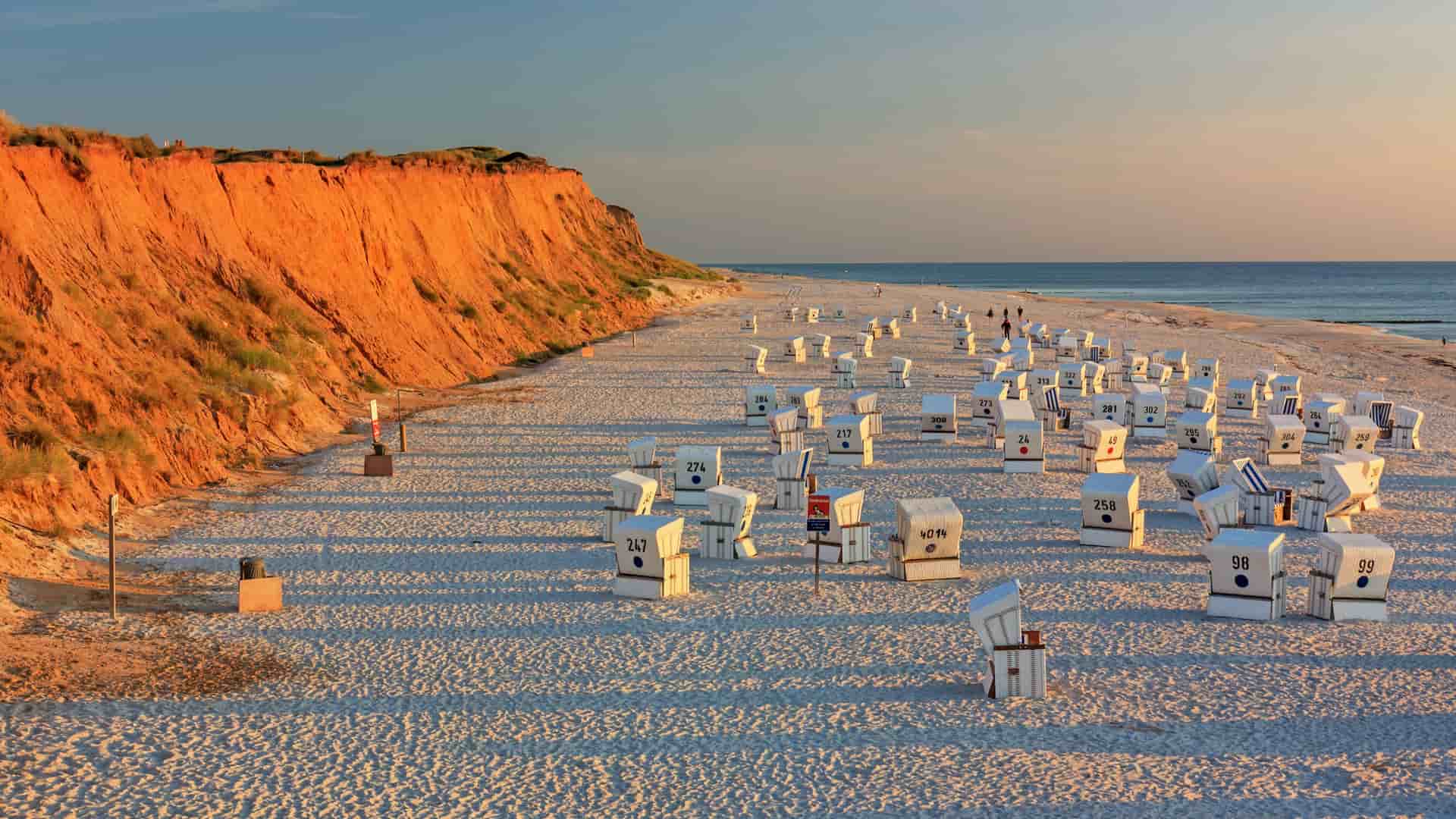 A serene sunset view of the Red Cliff beach in Kampen, on the island of Sylt, Germany, with numerous striped wicker beach chairs scattered on the sand and the calm North Sea in the background.