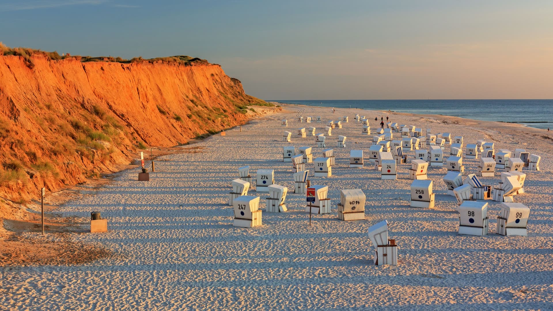 A serene sunset view of the Red Cliff beach in Kampen, on the island of Sylt, Germany, with numerous striped wicker beach chairs scattered on the sand and the calm North Sea in the background.