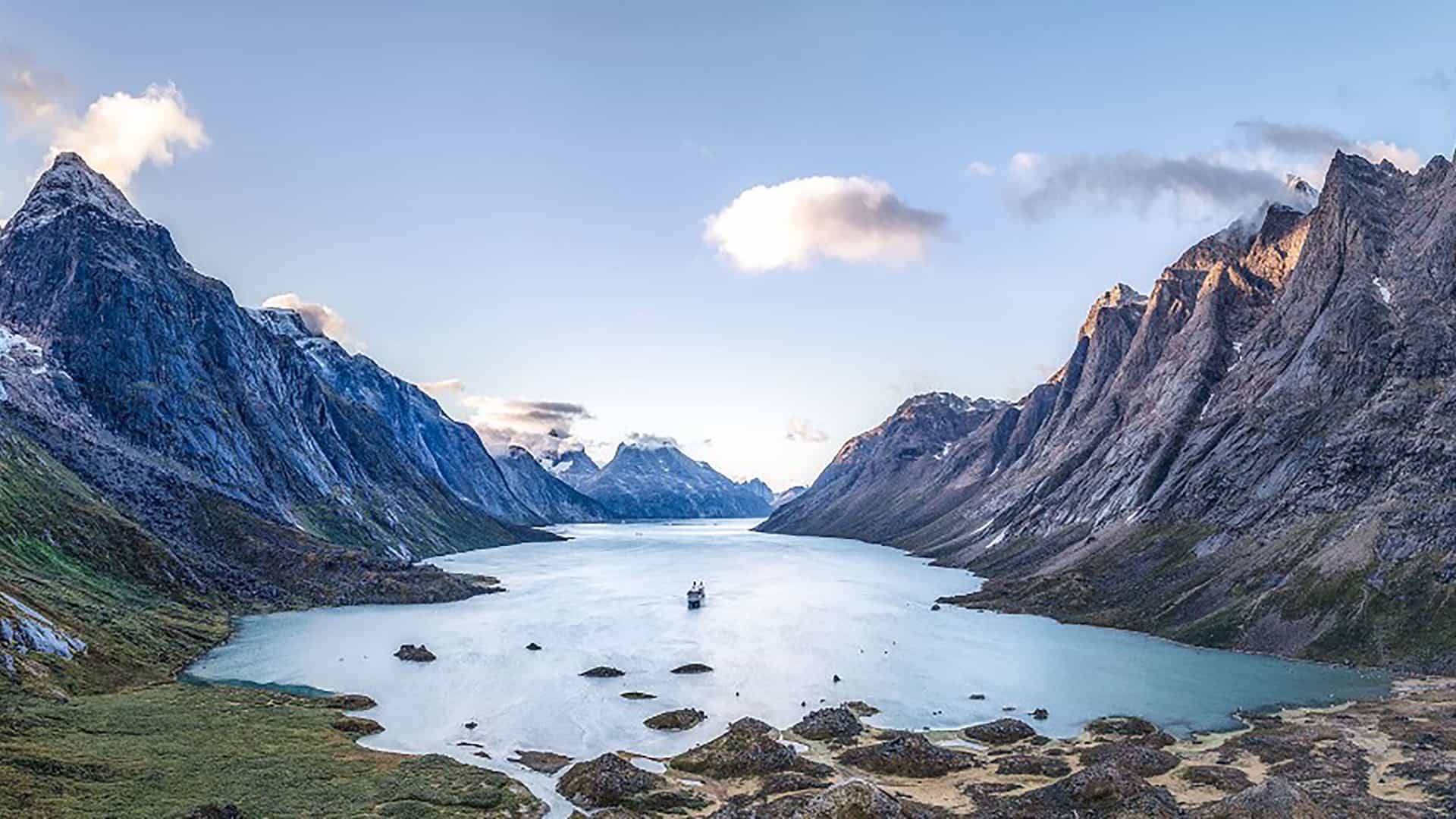 "A breathtaking panoramic shot of the stunning Tasermiut Fjord in Greenland, with a small ship sailing through the turquoise waters, surrounded by towering, snow-capped mountains under a wide blue sky. "