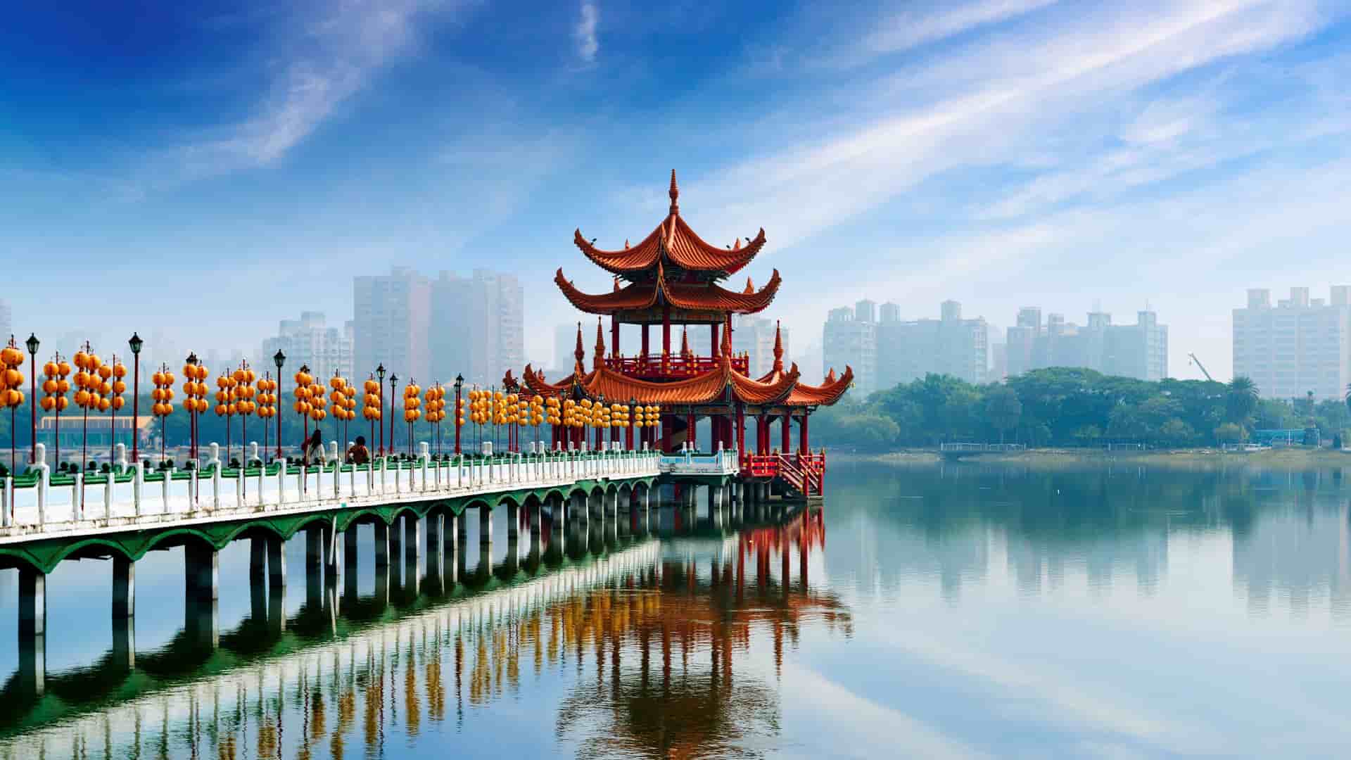 A picturesque view of the Spring and Autumn Pavilions at Lotus Lake in Kaohsiung, Taiwan, featuring a vibrant red traditional Chinese pavilion reflected in the water, with city buildings in the hazy background.