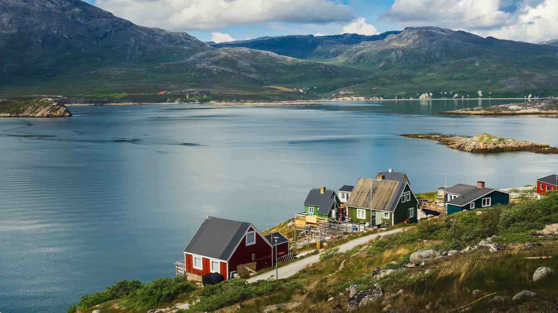 A panoramic view of the remote Kapisillit settlement in Greenland, with colorful houses situated on a grassy slope overlooking the serene fjord and majestic, rugged mountains in the distance.