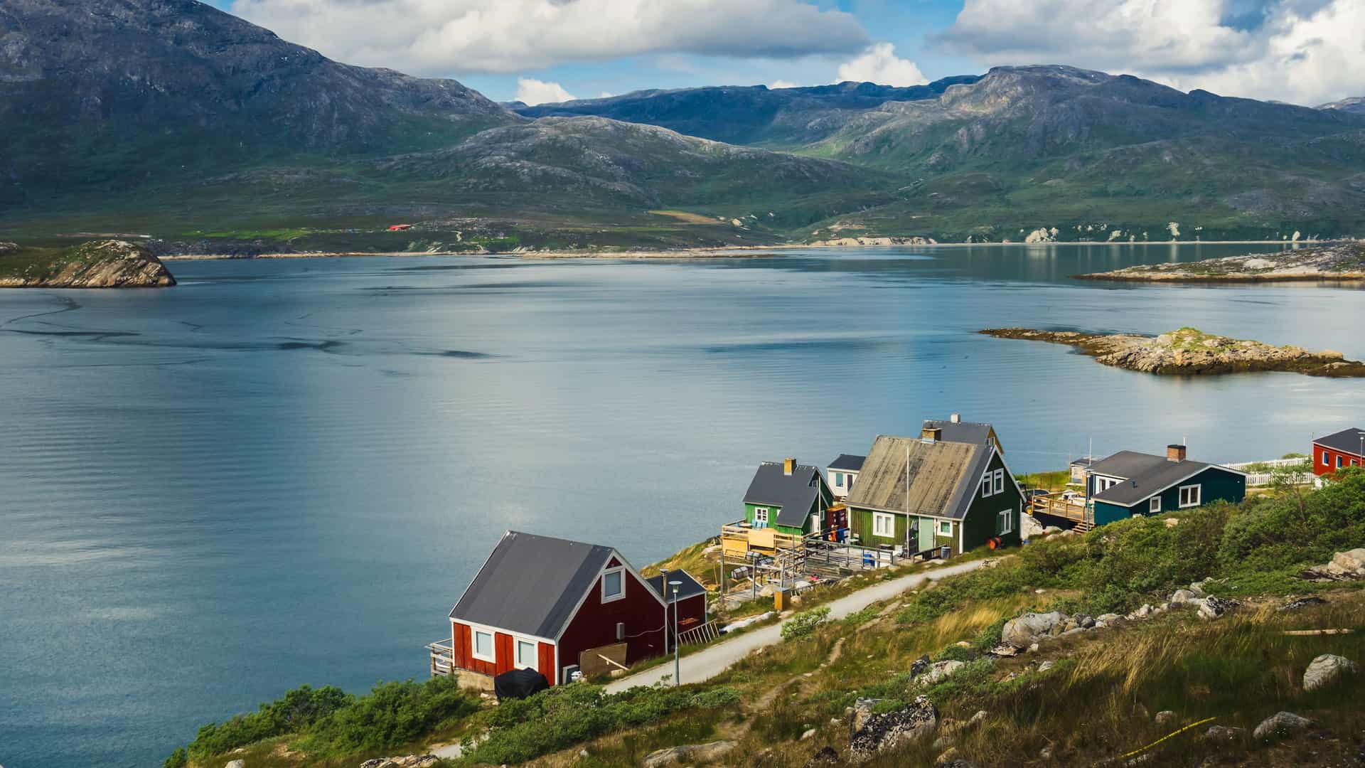 A panoramic view of the remote Kapisillit settlement in Greenland, with colorful houses situated on a grassy slope overlooking the serene fjord and majestic, rugged mountains in the distance.