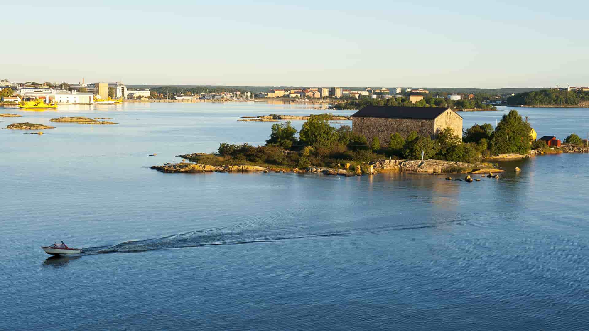 A tranquil landscape of the Karlskrona archipelago in Sweden, featuring a small island with a historic stone building and trees in the foreground, and the modern city skyline across the calm Baltic Sea in the distance.