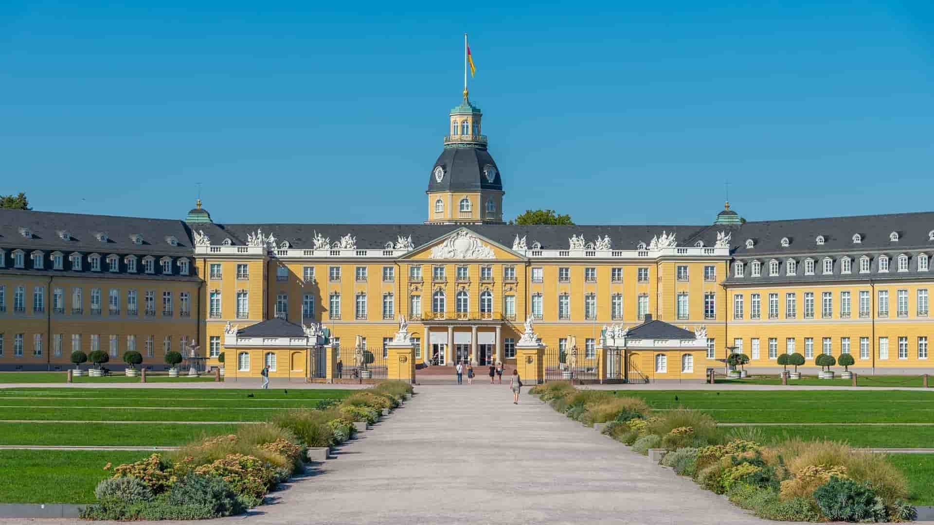 A beautiful daytime view of the Karlsruhe Palace, a grand yellow baroque building with a central tower, situated at the end of a long path with manicured green lawns and flowerbeds in Germany.