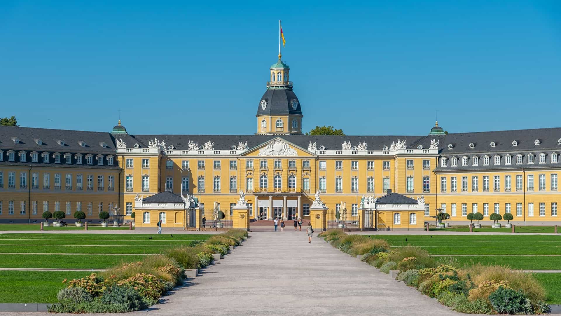 A beautiful daytime view of the Karlsruhe Palace, a grand yellow baroque building with a central tower, situated at the end of a long path with manicured green lawns and flowerbeds in Germany.