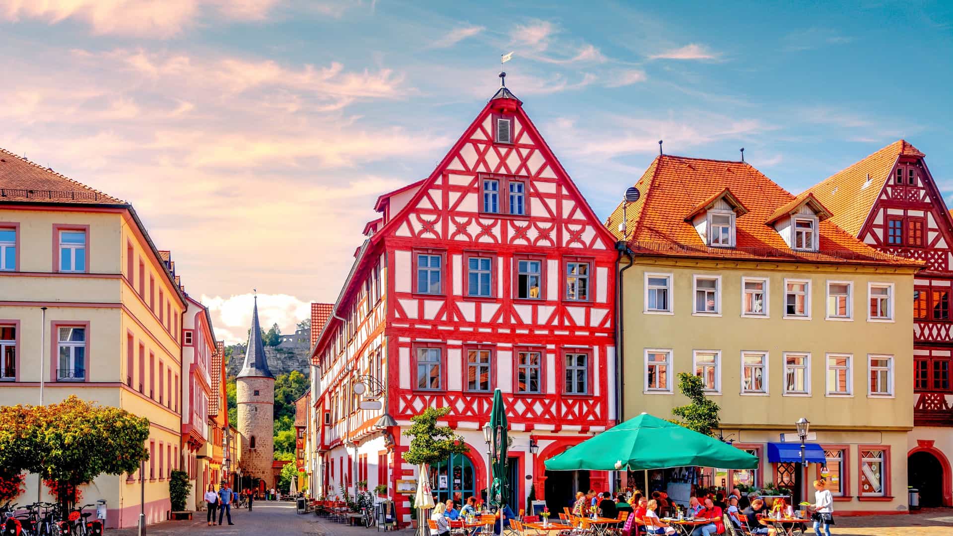 A bustling European square in Karlstadt am Main, Germany, with colorful half-timbered houses, outdoor cafes, and a distant church spire.