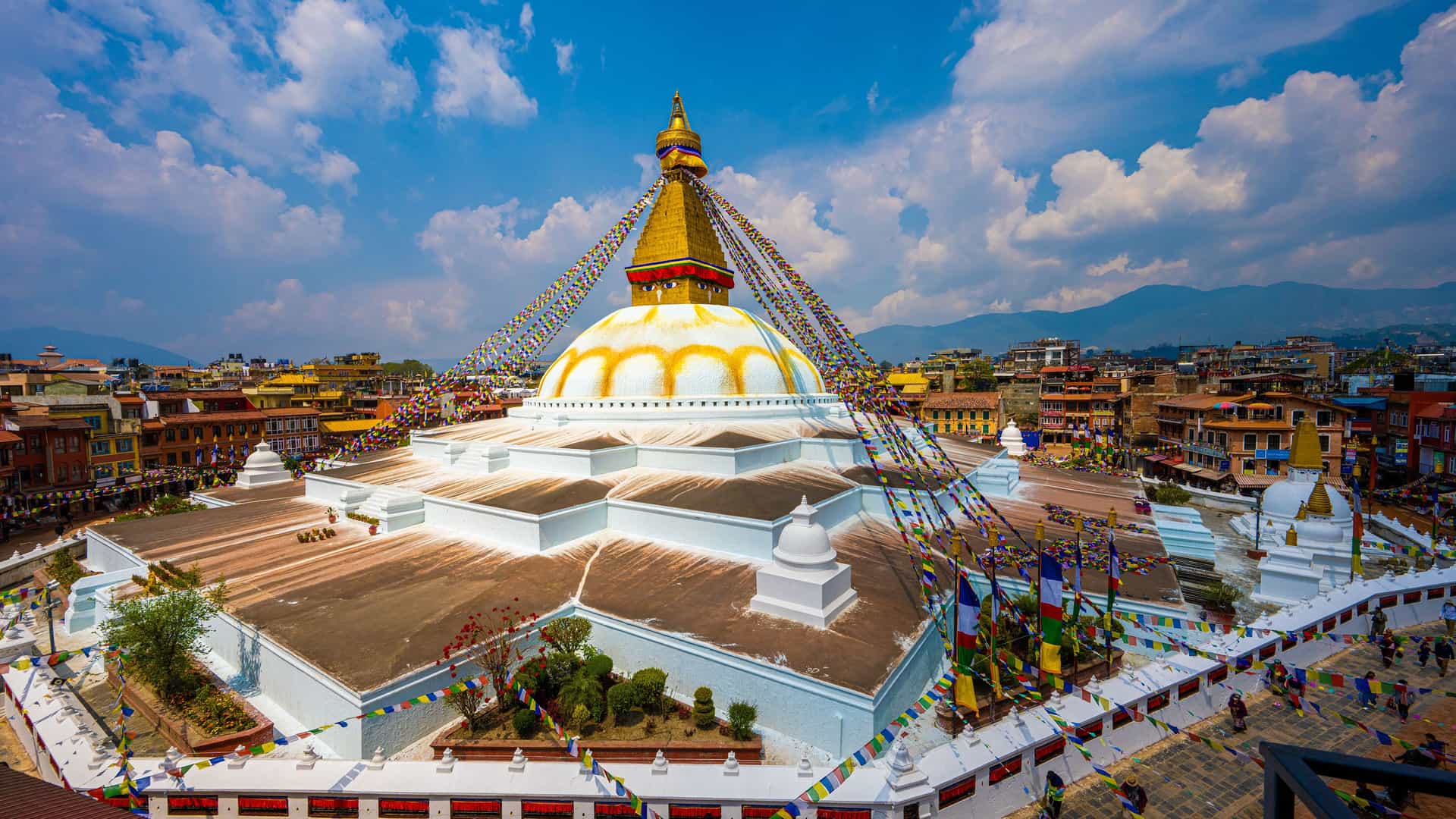 "A high-angle view of the Boudhanath Stupa, a large white-domed Buddhist monument in Kathmandu, Nepal, adorned with colorful prayer flags and a gilded spire under a blue sky. "