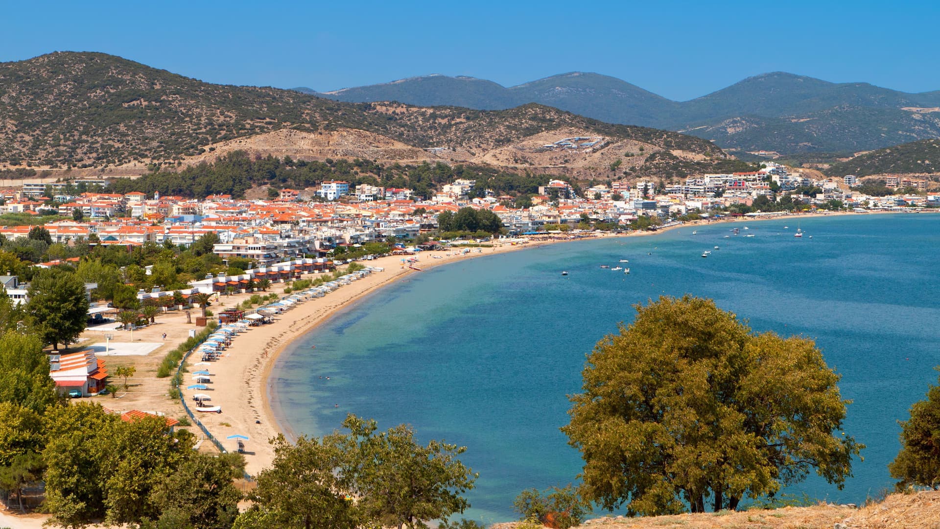A beautiful sunny view of Kavala, Greece, showing the curving beach, the town's buildings along the waterfront, and surrounding green hills.
