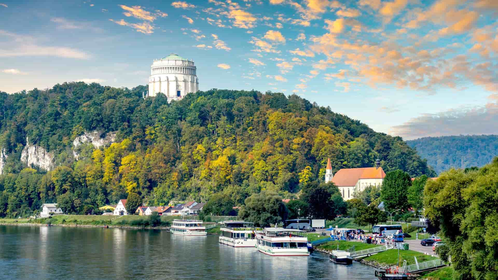 A scenic view of the town of Kelheim, Bavaria, Germany, showing tour boats docked on the wide, placid river. Above the town, the large, cylindrical Befreiungshalle (Hall of Liberation) monument sits prominently atop a forested hill under a dramatic sunset sky.