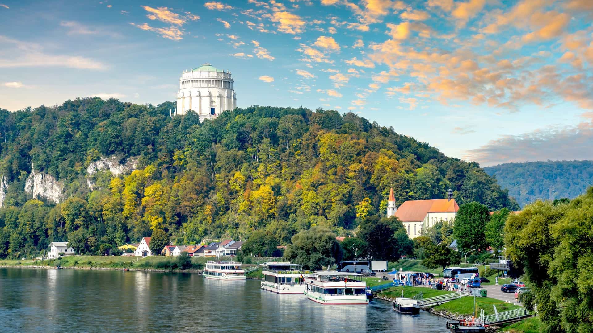 A scenic view of the town of Kelheim, Bavaria, Germany, showing tour boats docked on the wide, placid river. Above the town, the large, cylindrical Befreiungshalle (Hall of Liberation) monument sits prominently atop a forested hill under a dramatic sunset sky.