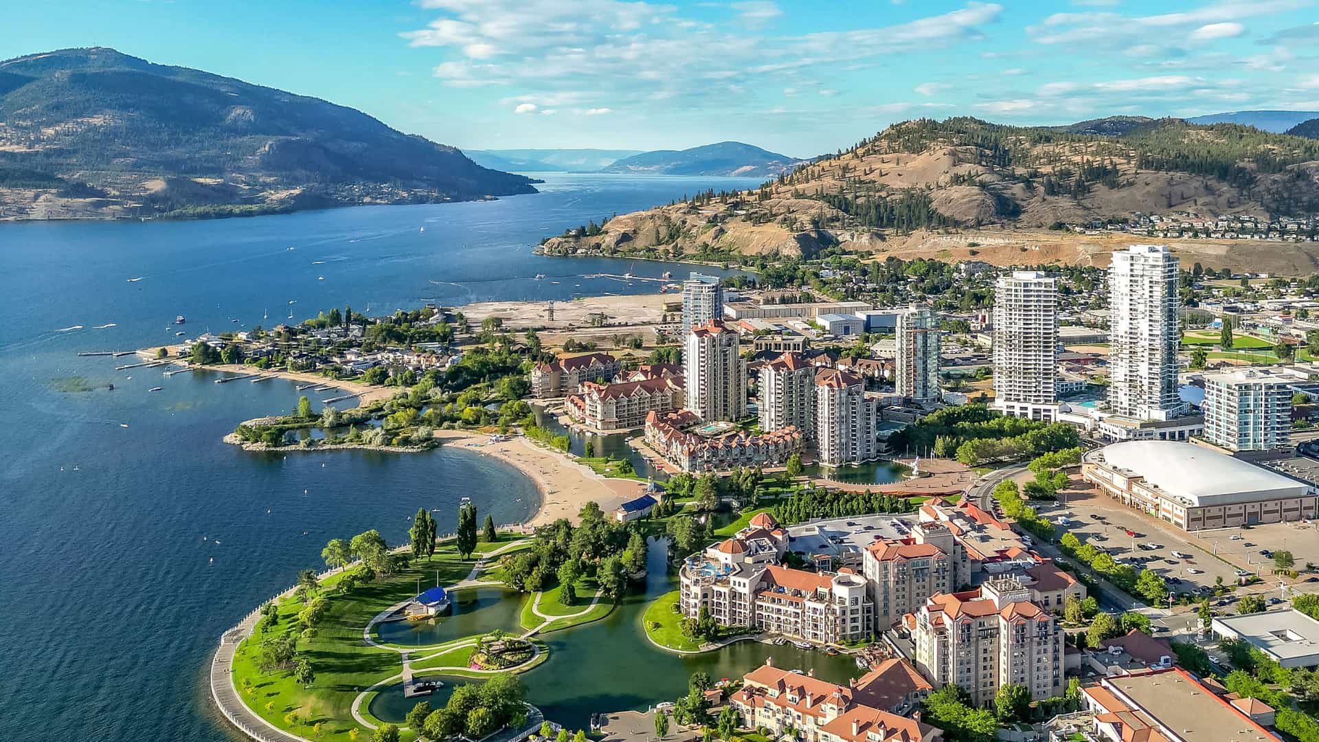 "An aerial shot of Kelowna, British Columbia, showing the city's waterfront and marina on the shores of Okanagan Lake, with modern high-rises and residential buildings against a backdrop of forested hills. "