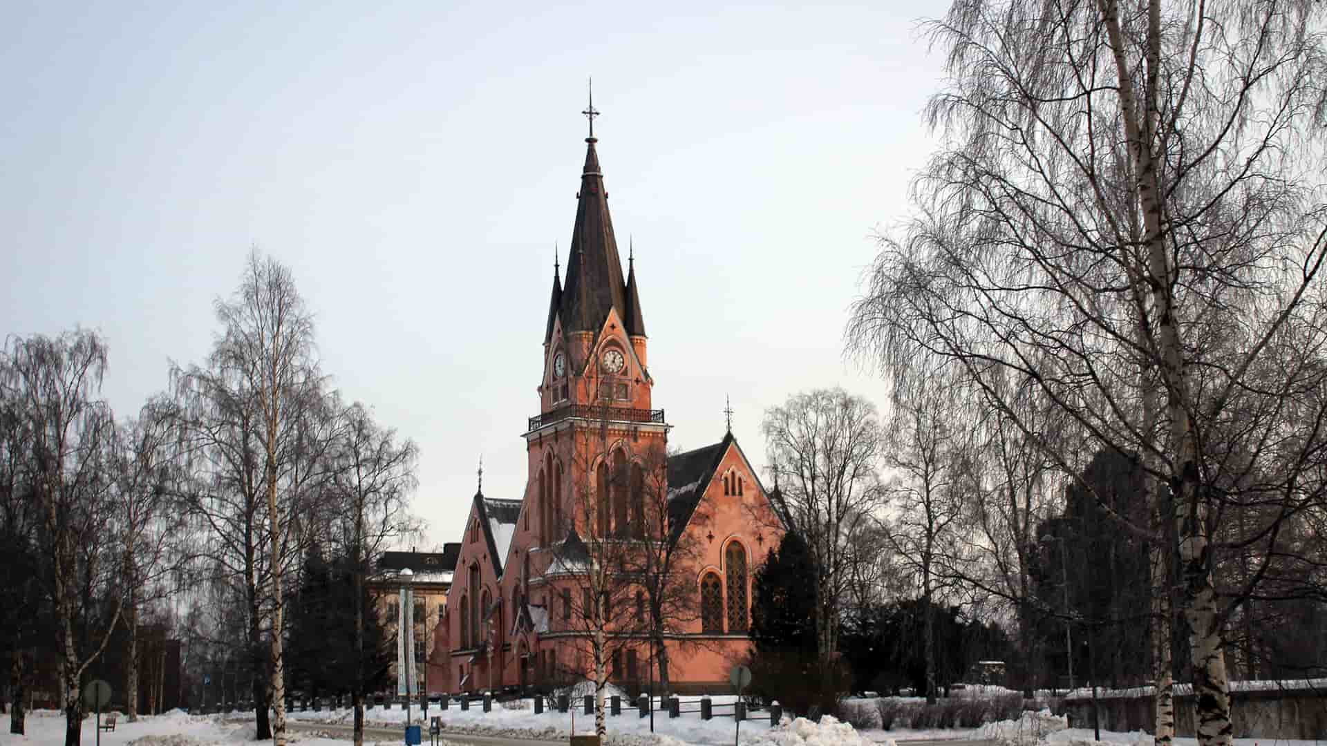 A pink-hued Kemi Church with a prominent steeple stands in a snowy Finnish landscape, surrounded by bare birch trees under a pale winter sky.