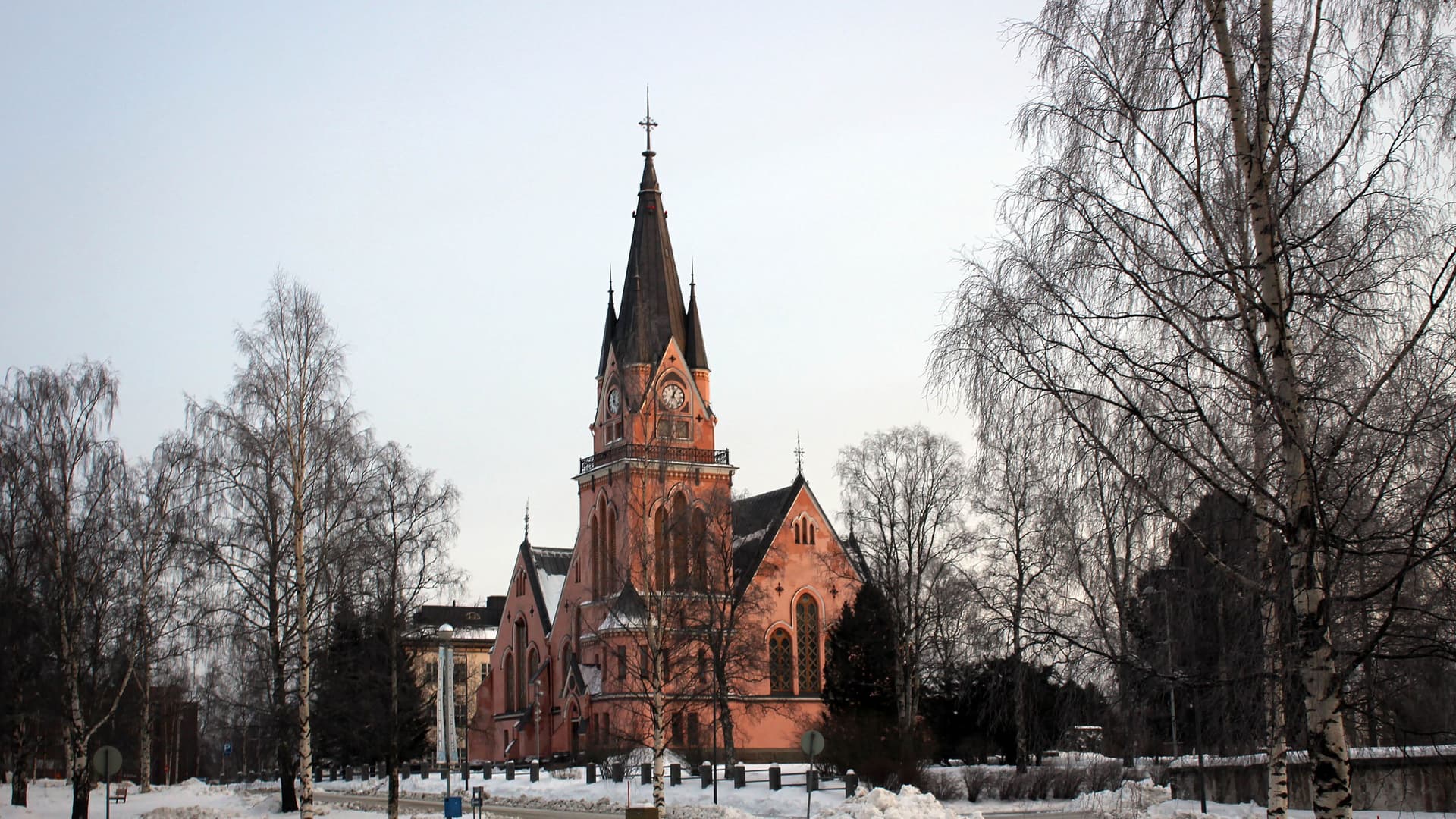 A pink-hued Kemi Church with a prominent steeple stands in a snowy Finnish landscape, surrounded by bare birch trees under a pale winter sky.