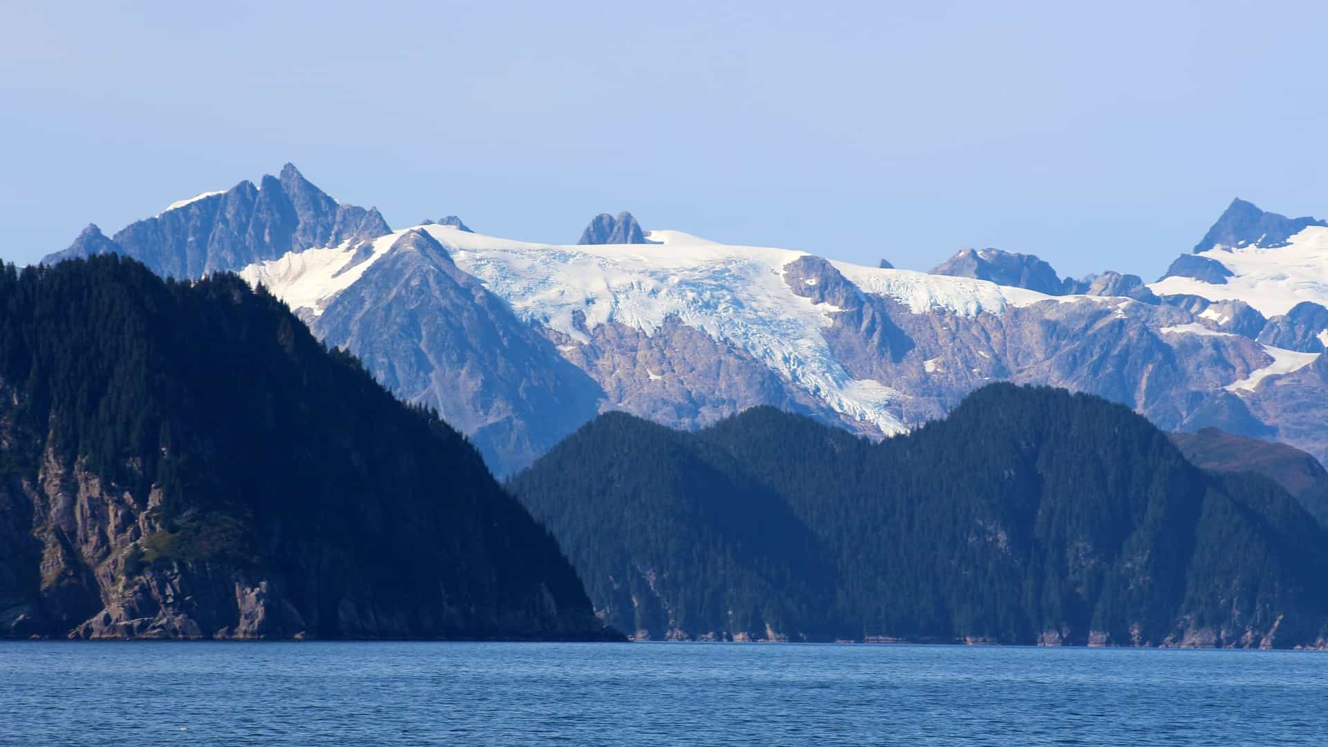 A dramatic view across the dark blue ocean water toward the Kenai Peninsula, Alaska, USA, featuring a series of steep, dark green forested coastal mountains that give way to towering peaks capped with bright white and blue glaciers.