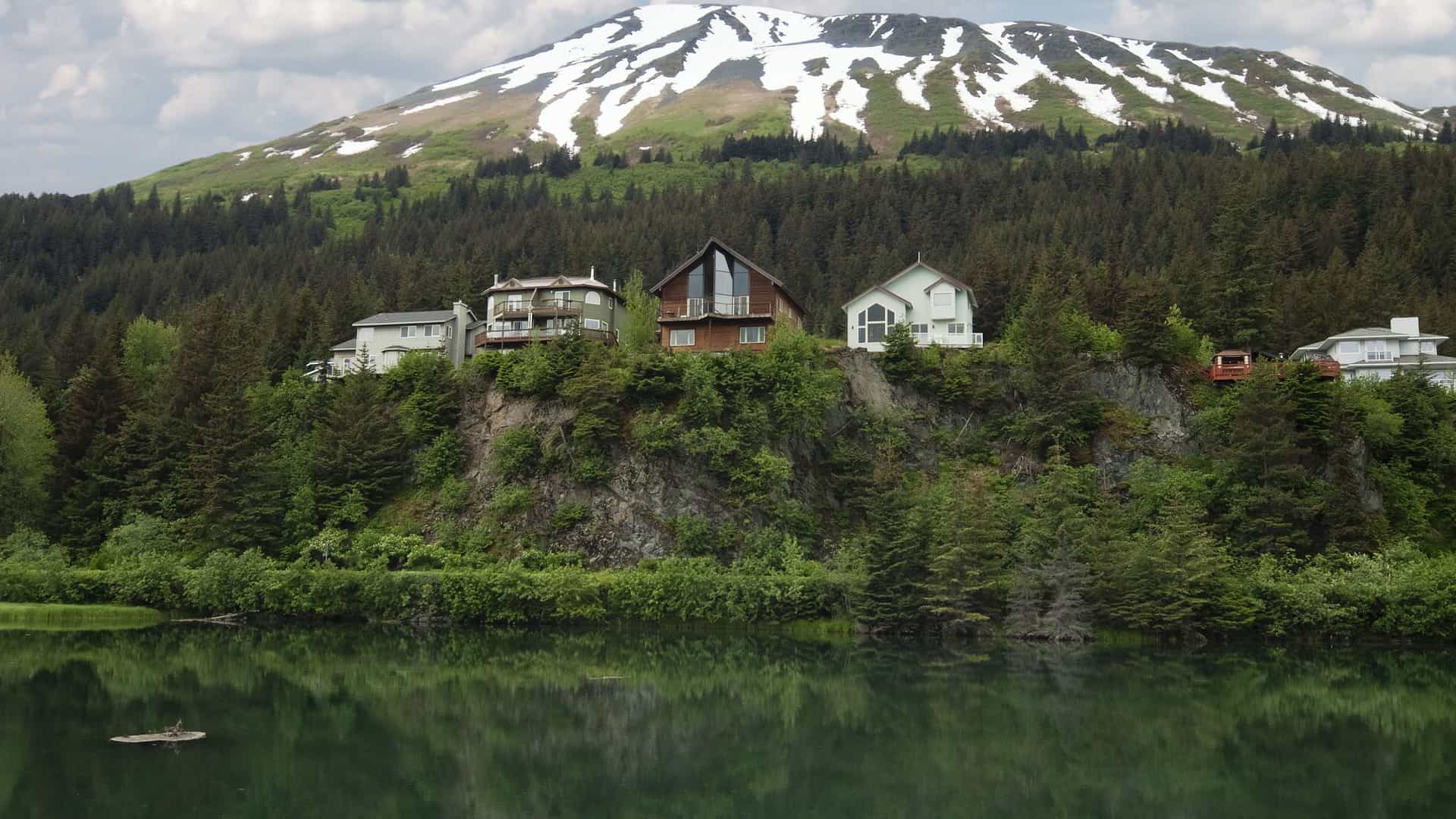 A beautiful waterfront view of the Kenai Princess Wilderness Lodge nestled on a cliffside, surrounded by thick forest with a snow-capped mountain in the background.