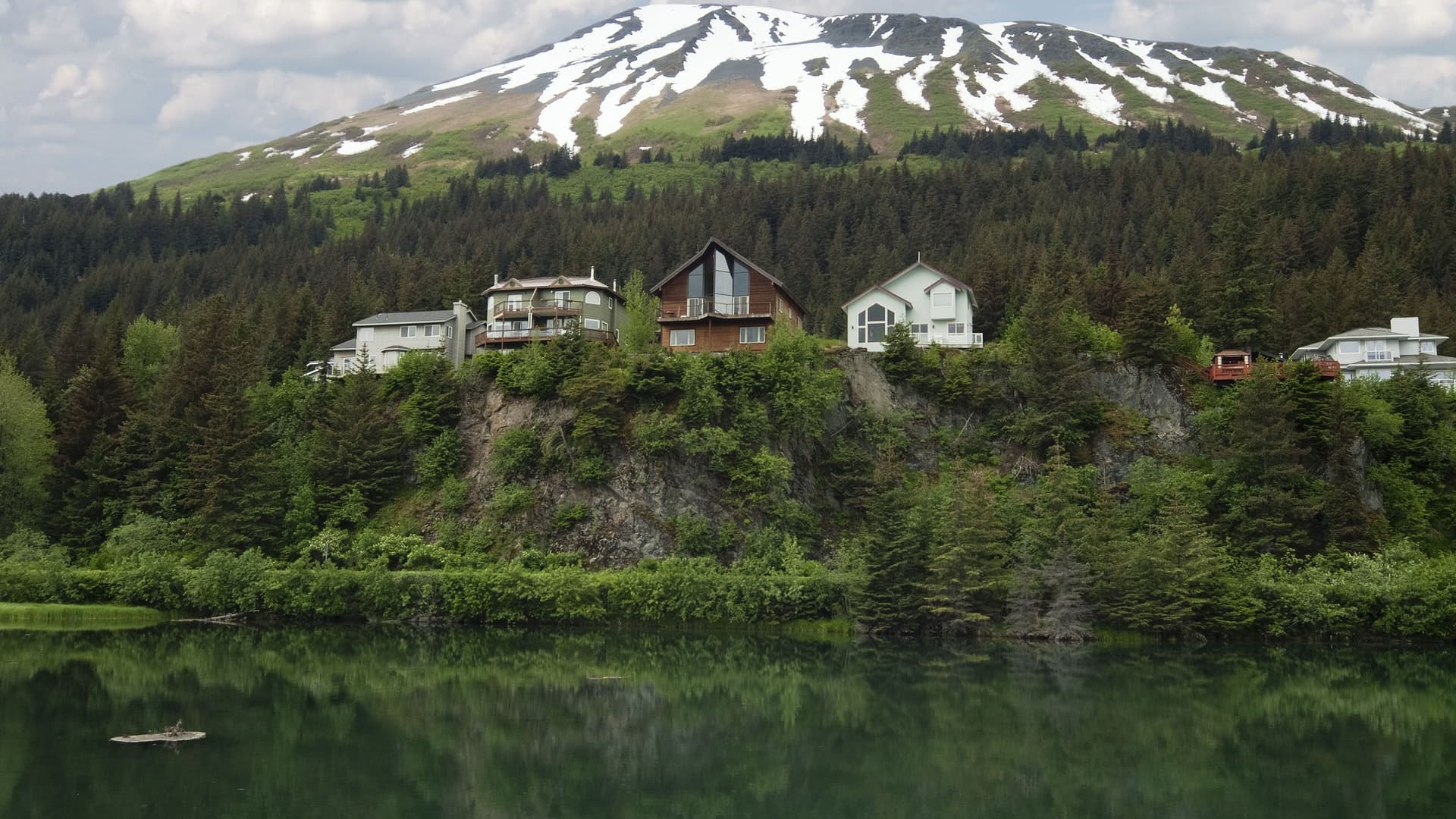 A beautiful waterfront view of the Kenai Princess Wilderness Lodge nestled on a cliffside, surrounded by thick forest with a snow-capped mountain in the background.