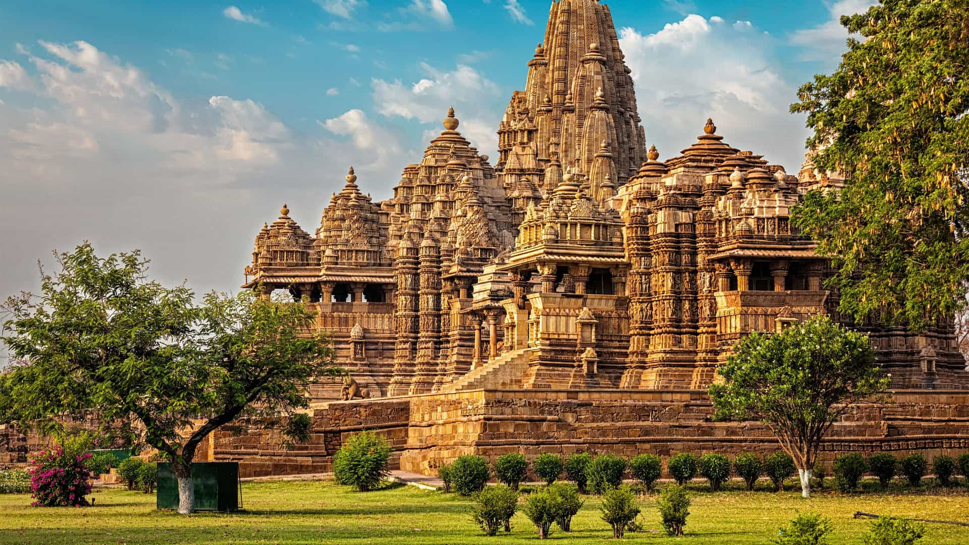 "A magnificent view of the Kandariya Mahadeva Temple, the largest of the ancient Hindu temples in Khajuraho, India, with its intricately carved spires and detailed stone architecture, surrounded by green lawns and trees. "