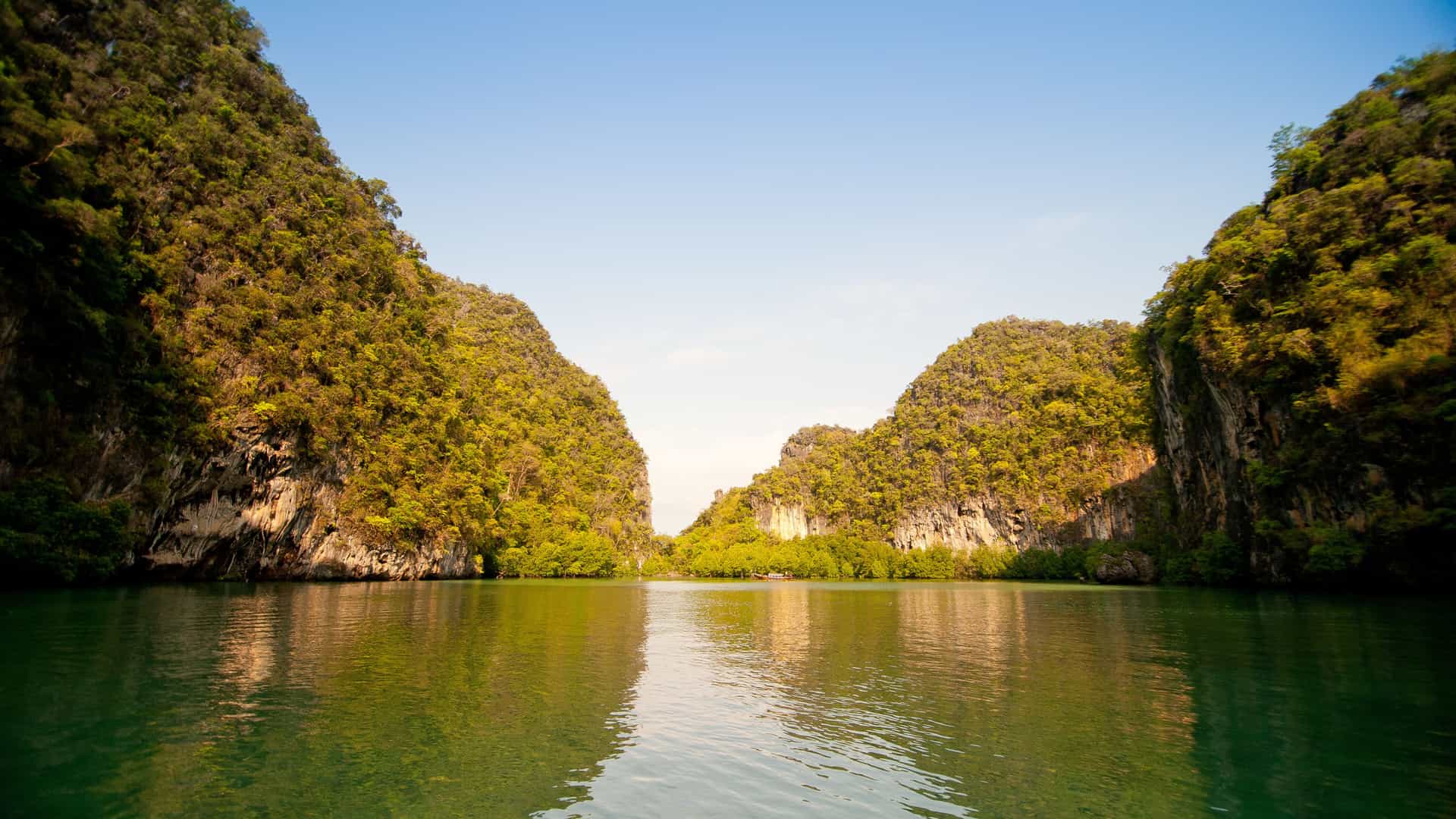 A tranquil tropical scene in Kho Yao with calm reflective water framed by steep forested limestone cliffs, forming a narrow passage beneath a clear light‑blue sky.