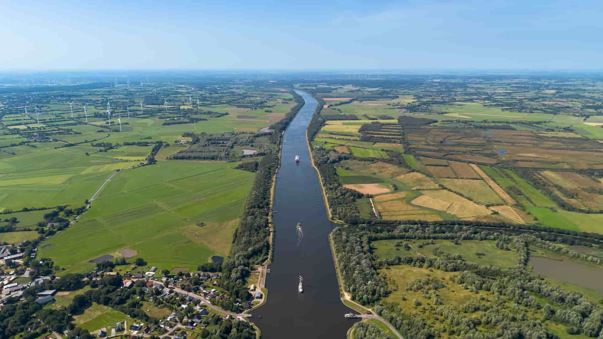 An aerial shot of the Kiel Canal in Germany, with multiple ships navigating the waterway surrounded by flat, green farmland and numerous wind turbines on a clear, sunny day.