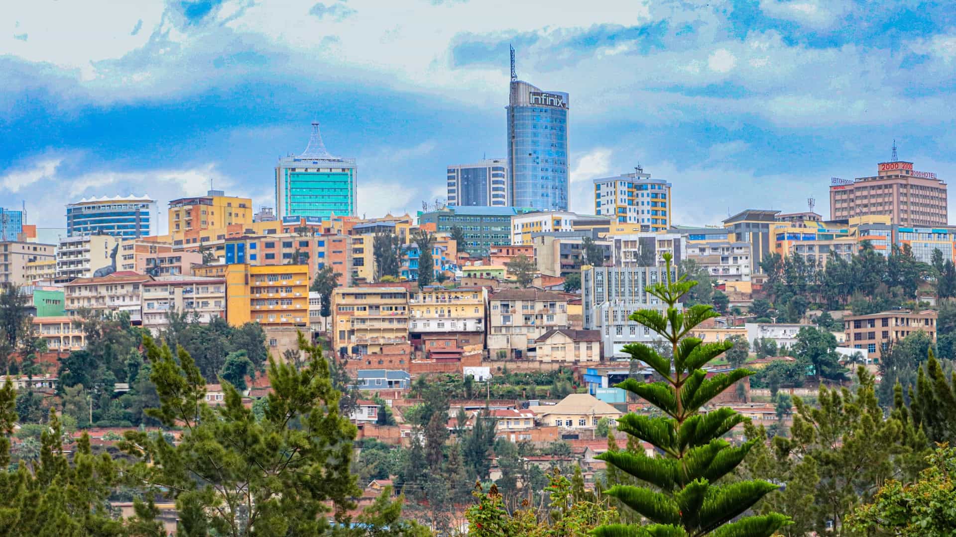 A dynamic view of the modern Kigali, Rwanda skyline, featuring several colorful buildings and high-rise towers, including the tall, cylindrical Radisson Blu Hotel on the hill, set against a bright, cloudy blue sky, with a foreground of green trees and foliage.