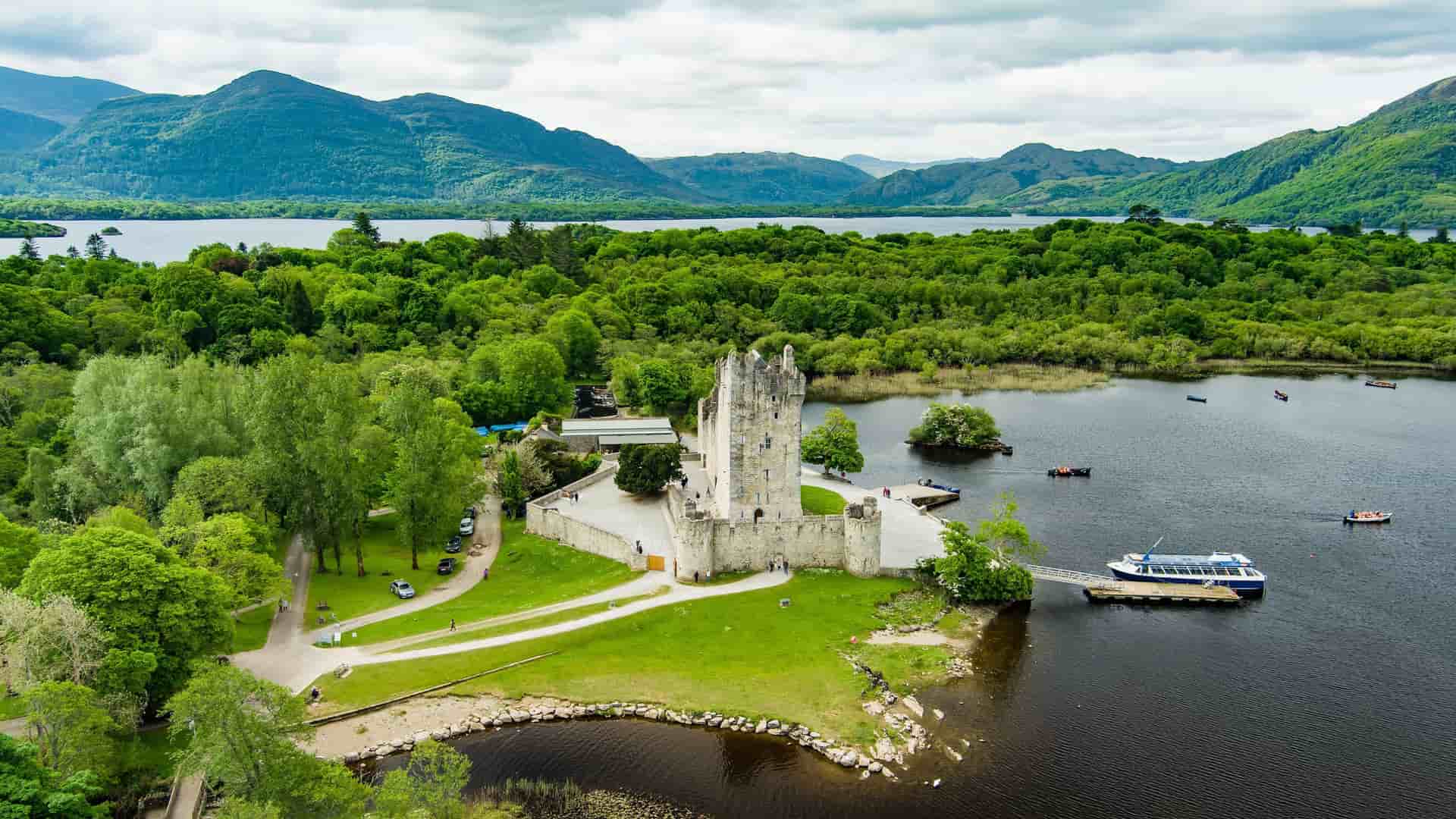 An aerial view of Ross Castle, a medieval fortress on the shores of Lough Leane, surrounded by lush green forests and mountains in Killarney National Park, Ireland.