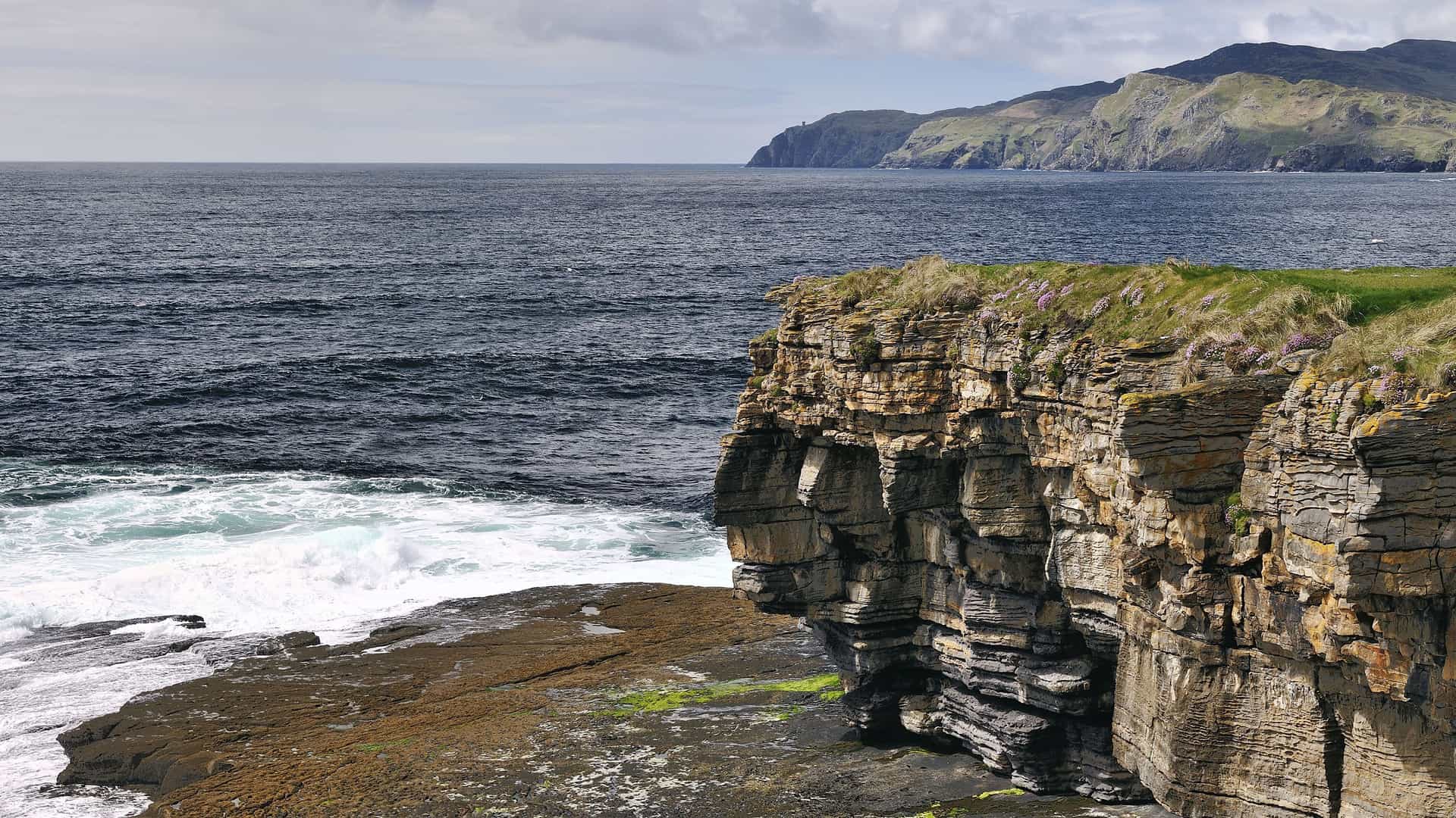 A scenic view of the rugged coastline of Killybegs, Ireland, with waves crashing against dramatic, layered rock cliffs and rolling green hills in the distance.