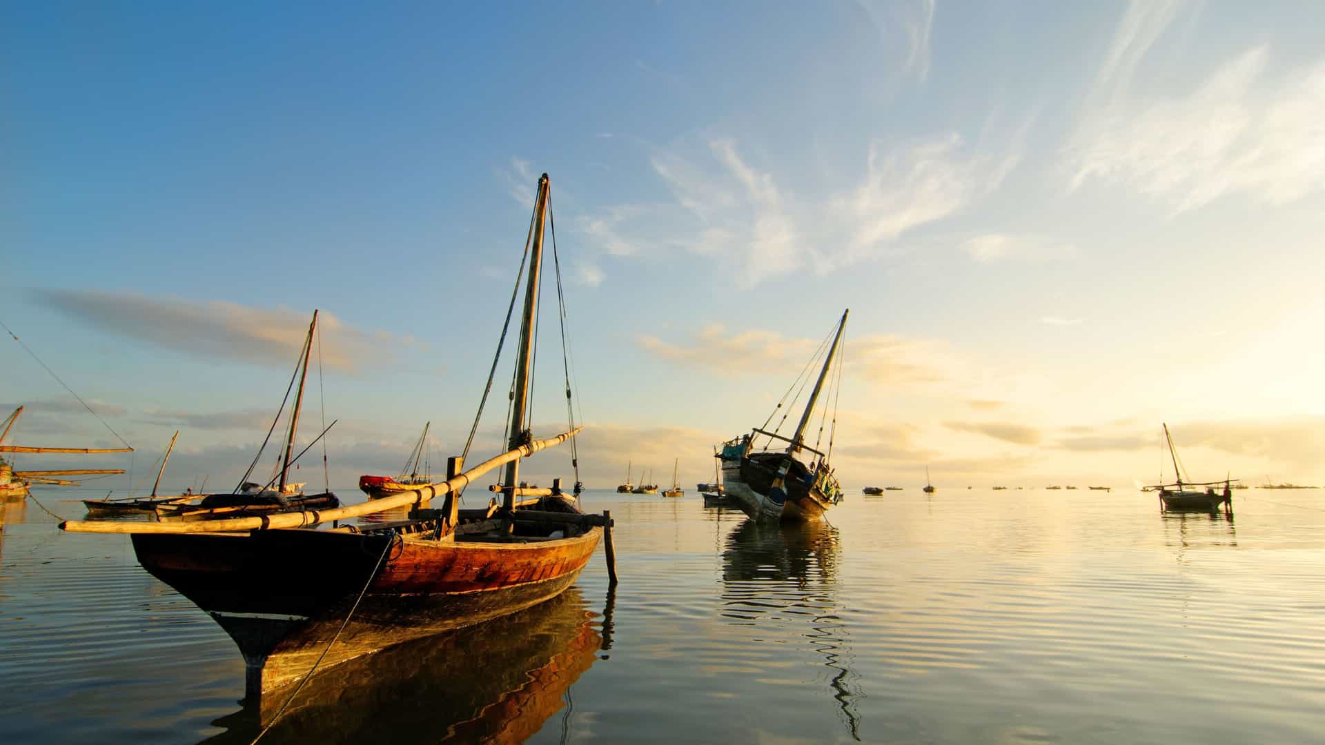 Traditional wooden dhow boats floating on calm water in the Indian Ocean near Kilwa Kisiwani, with a soft, hazy sunrise filling the sky with warm colors.