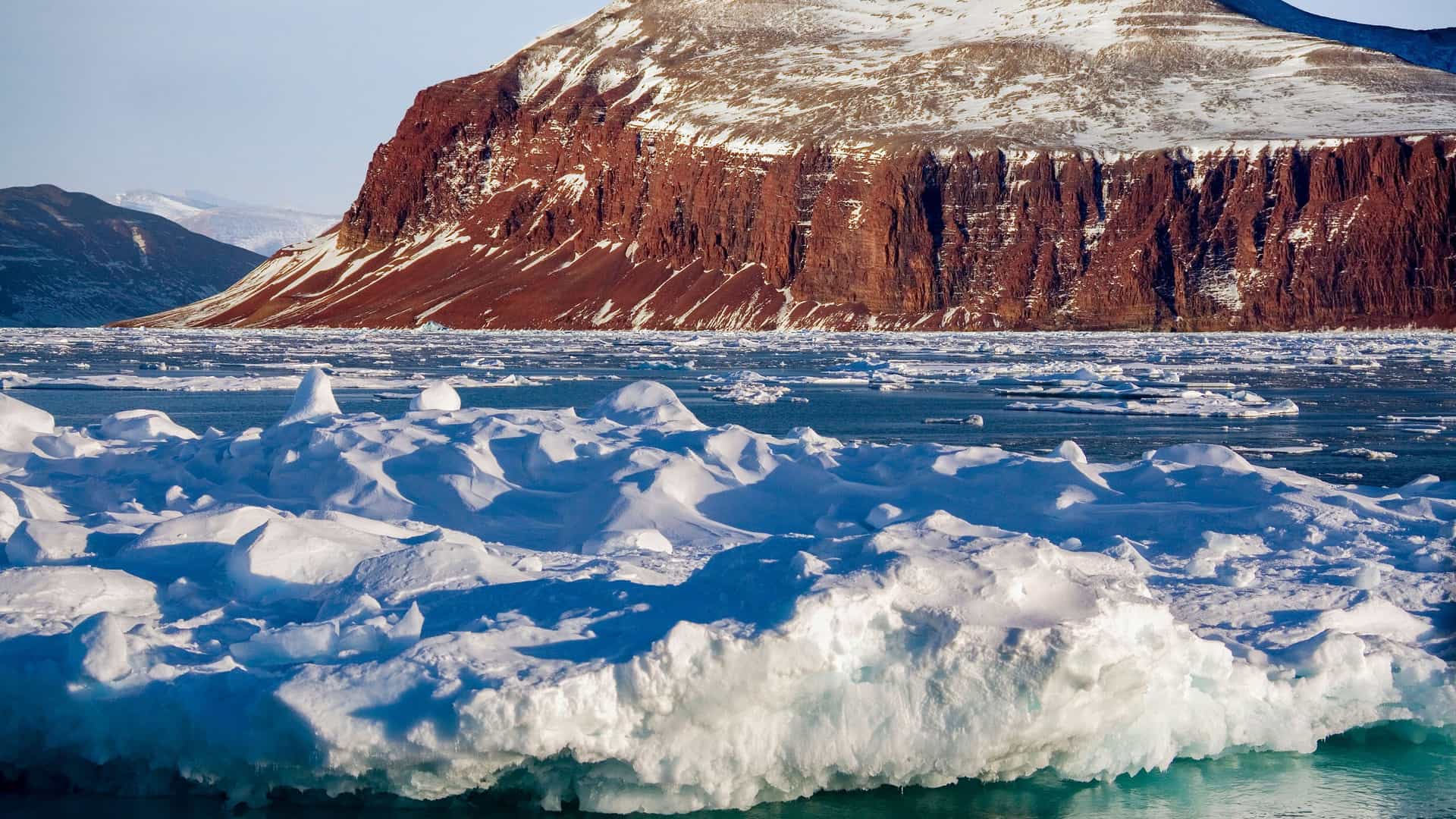 A close-up view of ice floes and large icebergs in the frigid, turquoise water of King Oscar Fjord in Greenland, with a dramatic red and white-striped mountain in the background.