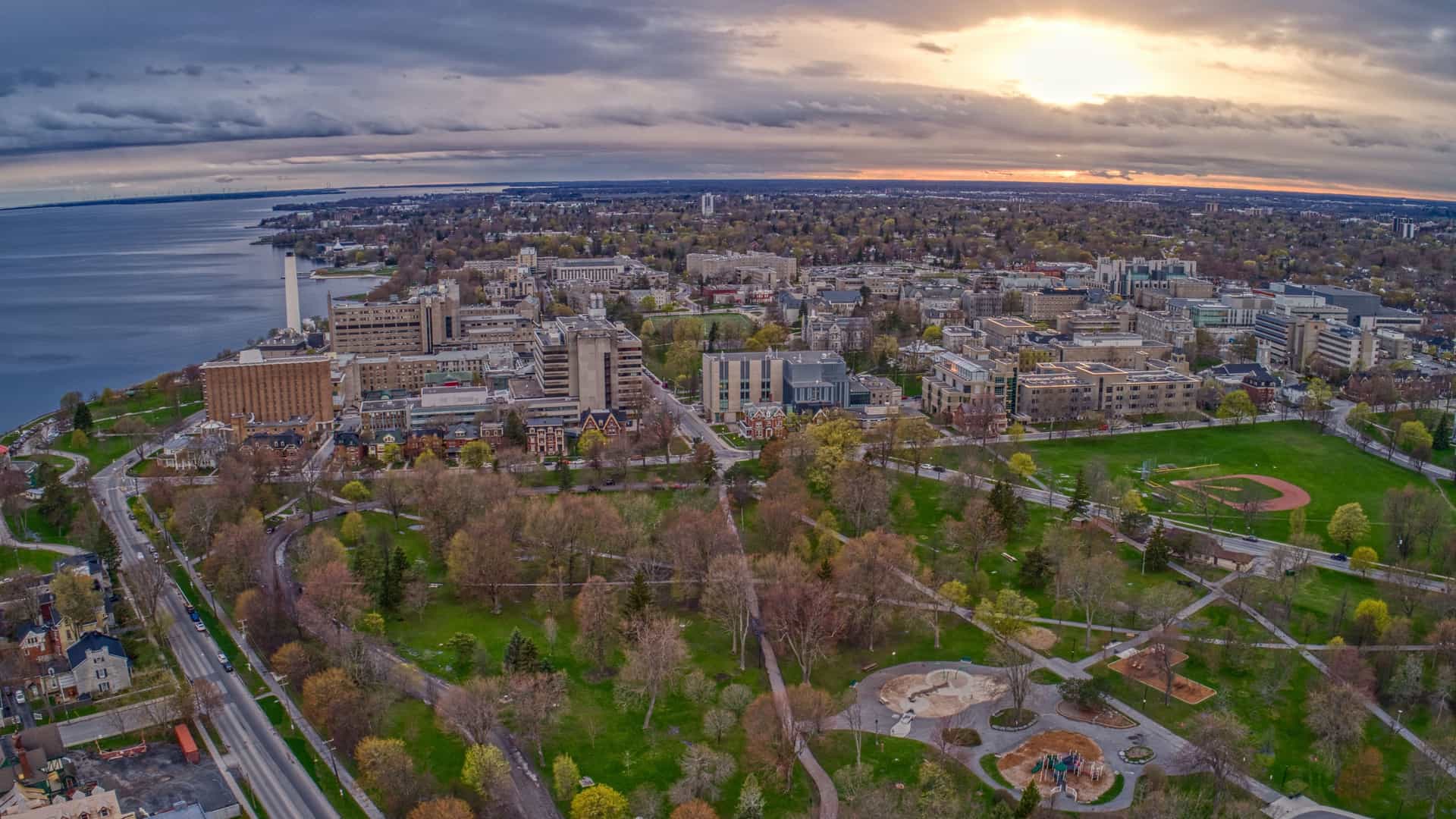 "An aerial panoramic shot of Kingston, Ontario, with the Cataraqui River and Lake Ontario on the left, showing a wide view of the city's parks, residential areas, and the Kingston General Hospital campus under a cloudy sky. "