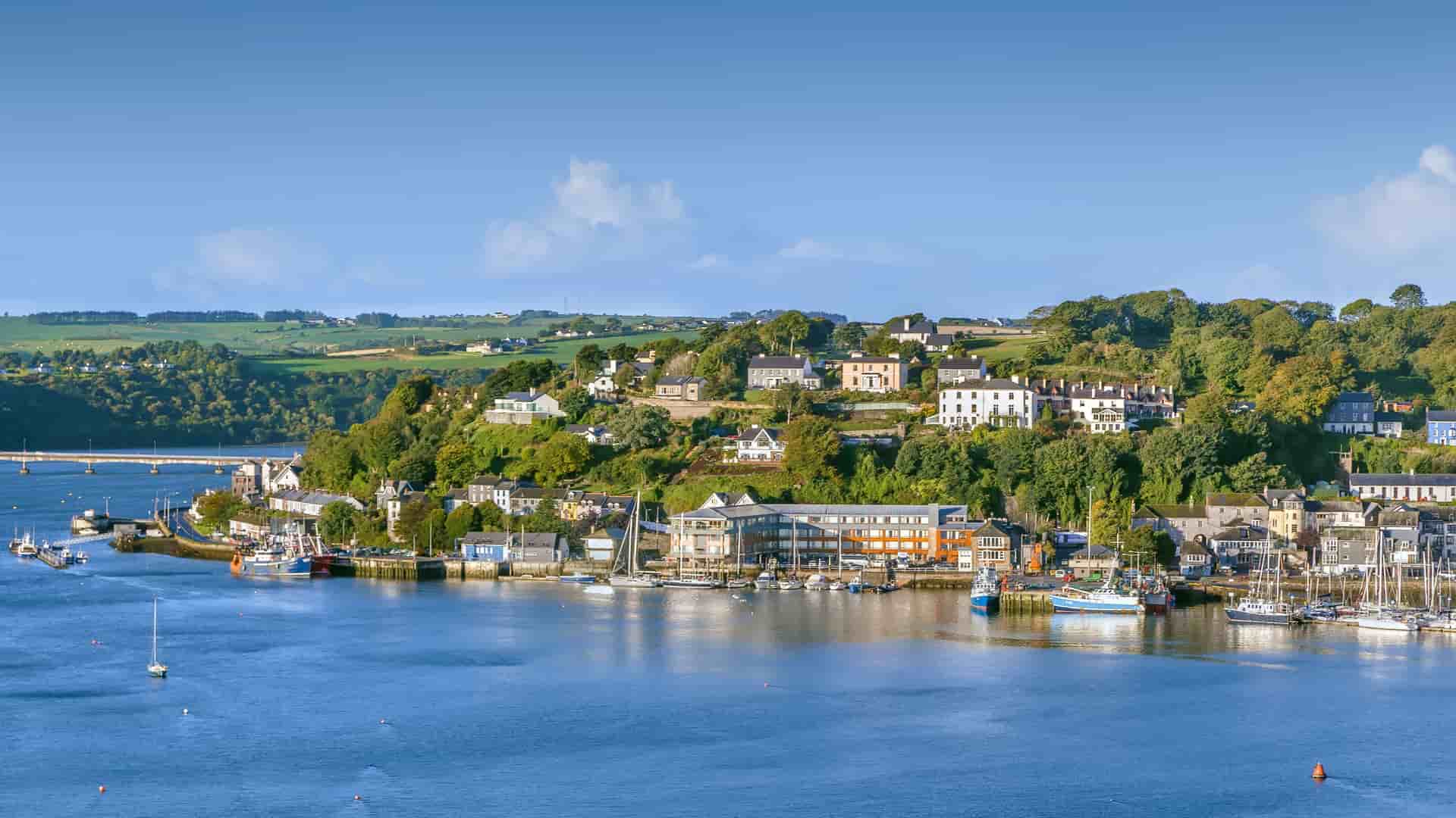 "A beautiful harbor shot of the coastal town of Kinsale, Ireland, with colorful buildings and houses lining the steep hills overlooking the calm water, with boats and yachts moored in the marina. "