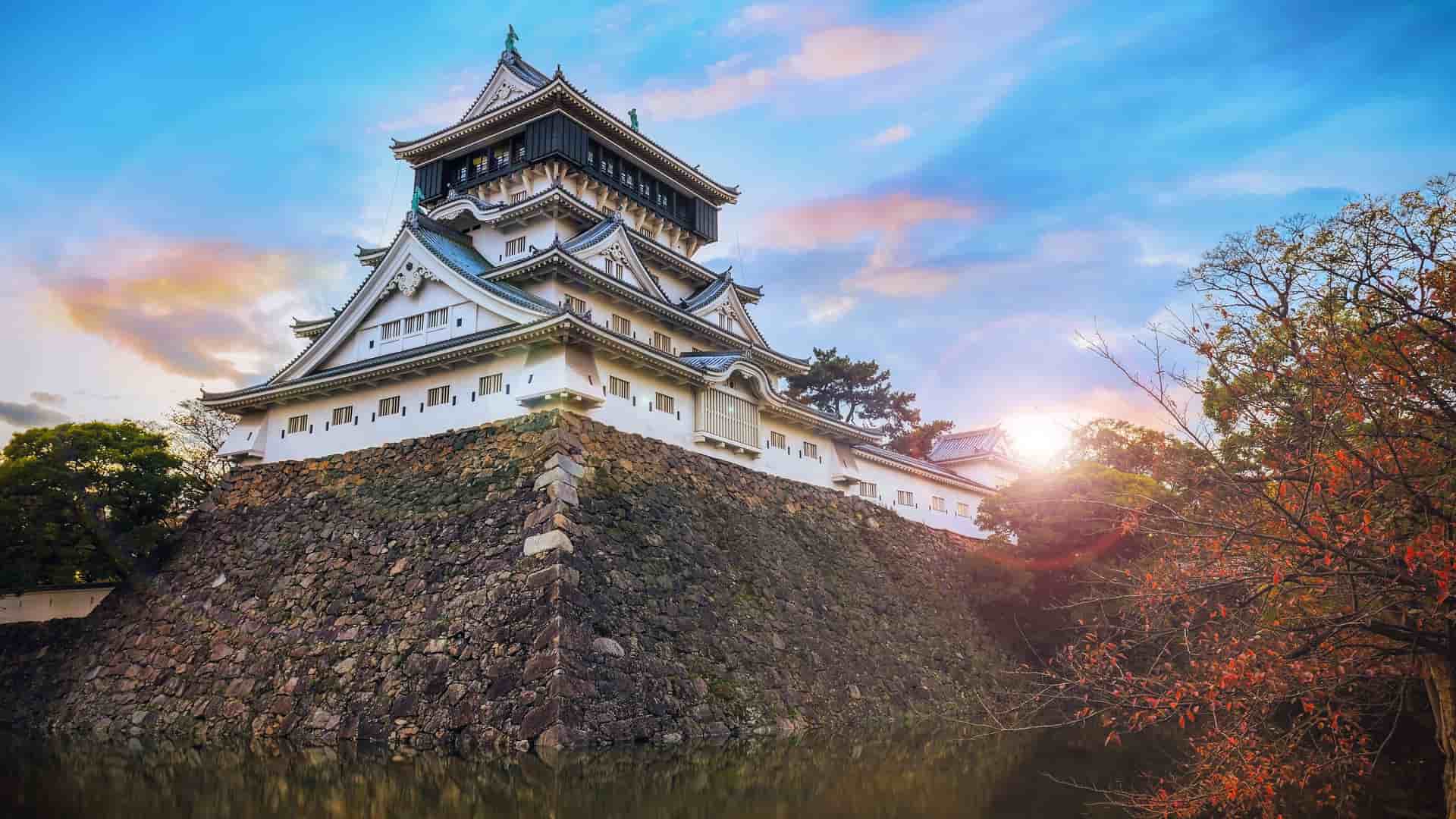 A majestic view of Kokura Castle in Kitakyushu, Japan, with its traditional white architecture and black roof, perched on a stone wall overlooking a moat at sunset.
