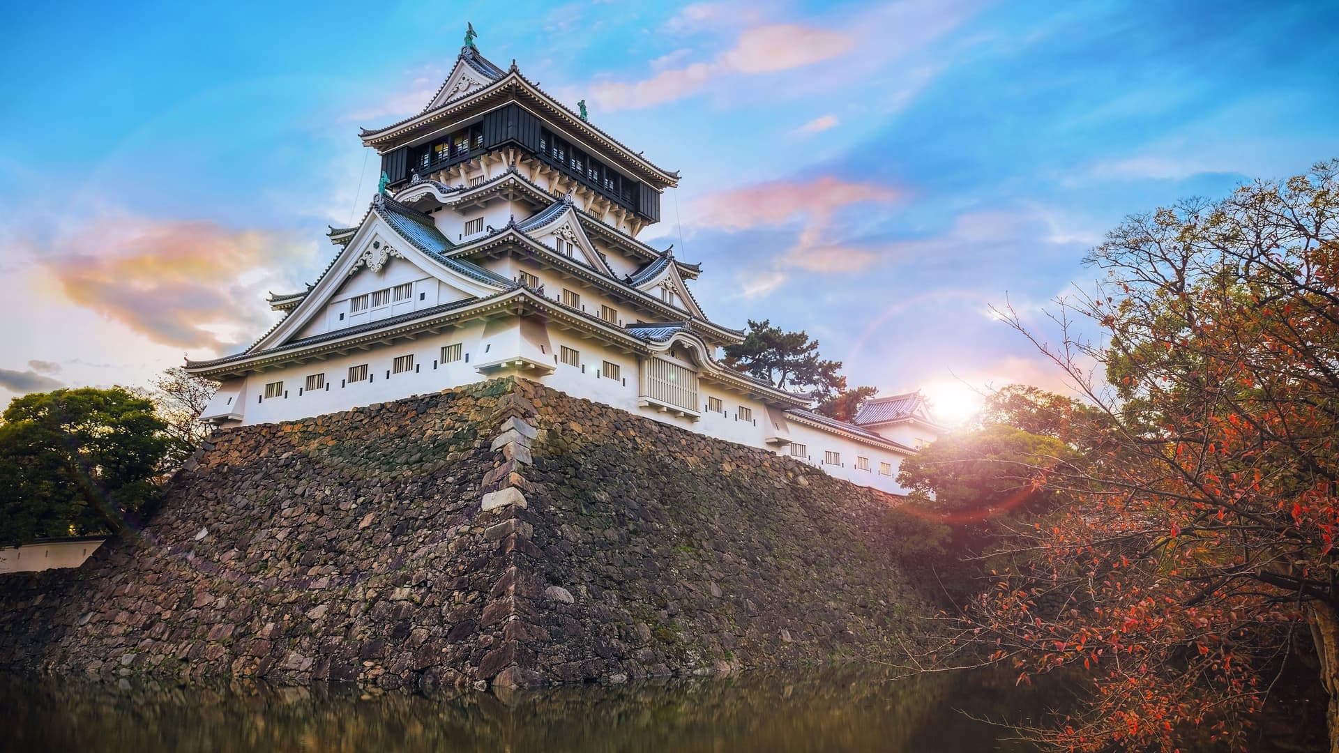 A majestic view of Kokura Castle in Kitakyushu, Japan, with its traditional white architecture and black roof, perched on a stone wall overlooking a moat at sunset.