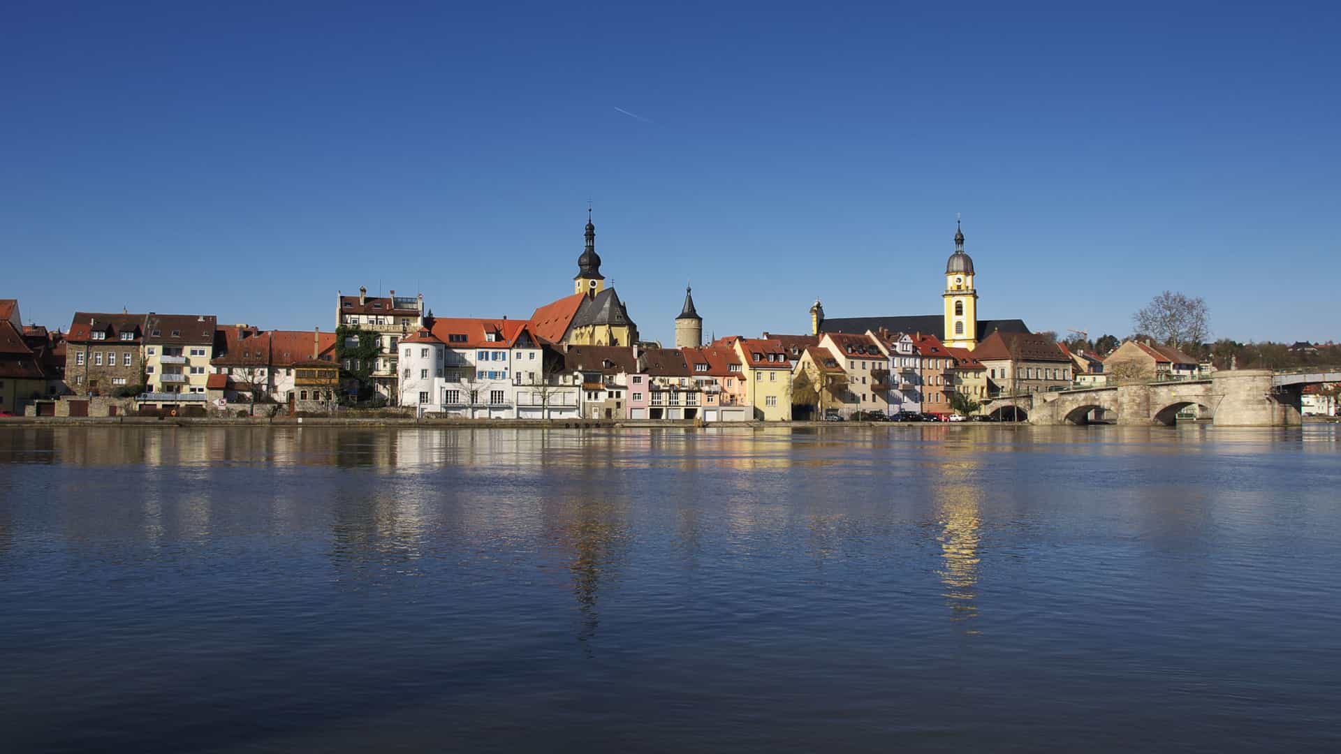 A serene panoramic view of the historic town of Kitzingen, Germany, with colorful buildings and church spires reflecting in the tranquil Main River under a clear blue sky.