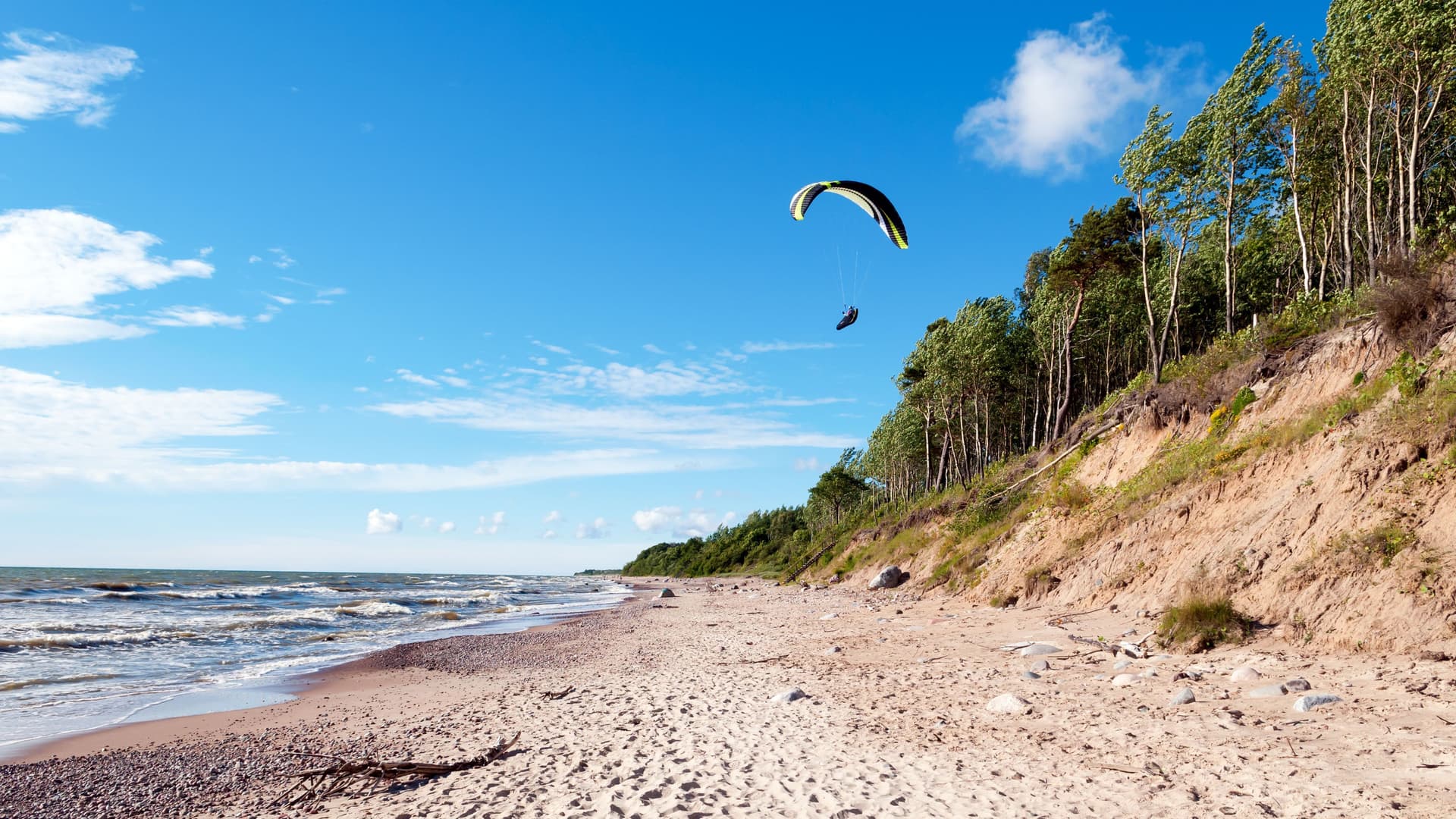 A person paragliding over a sandy beach near Klaipeda, Lithuania, with waves from the Baltic Sea crashing on the shore and a wooded cliffside under a vibrant blue sky with scattered clouds.