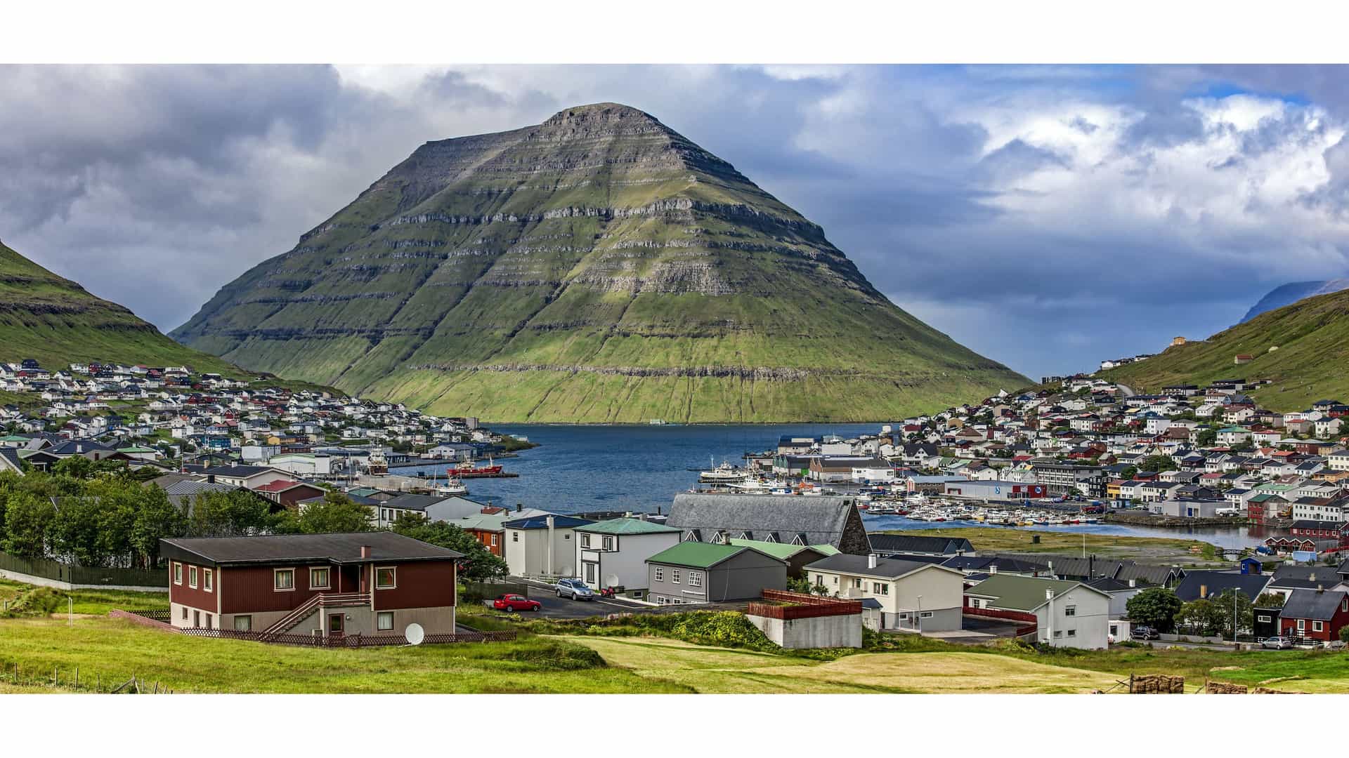 A scenic view of Klaksvík, the second-largest town in the Faroe Islands, nestled in a valley between green, conical mountains, with a harbor filled with boats.