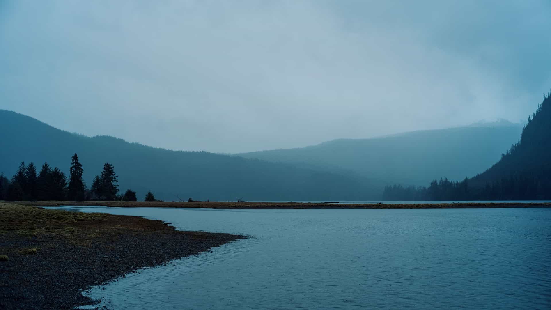 A tranquil, moody landscape of Klawock, Alaska, with a calm lake reflecting dark, forested mountains in the distance under an overcast and hazy sky.