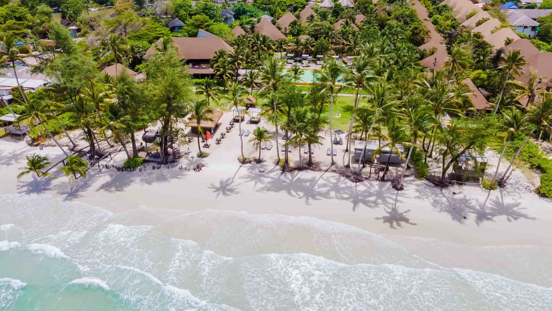 An aerial view of a luxurious resort on Ko Kut (Koh Kood), Thailand, with thatched-roof bungalows, palm trees, and lounge chairs on a white sandy beach with gentle waves.