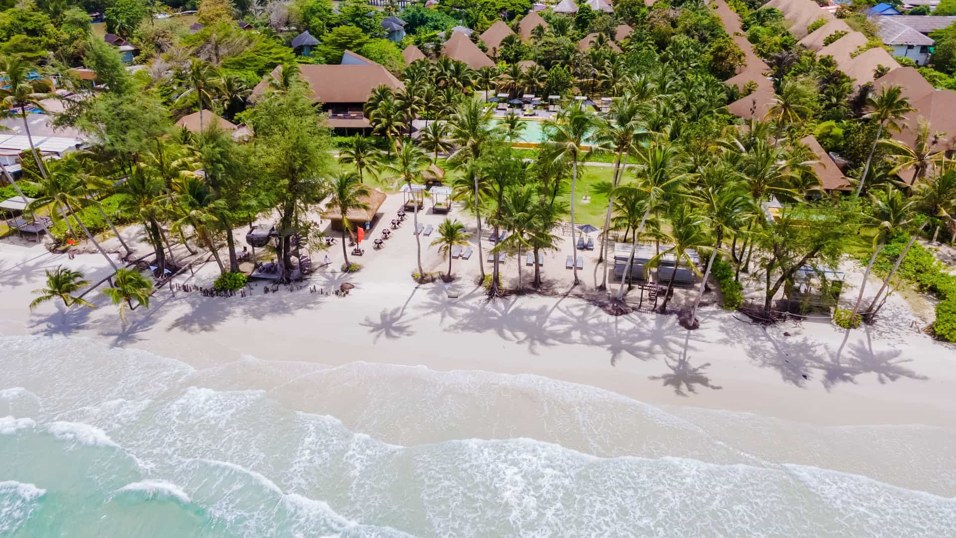 An aerial view of a luxurious resort on Ko Kut (Koh Kood), Thailand, with thatched-roof bungalows, palm trees, and lounge chairs on a white sandy beach with gentle waves.