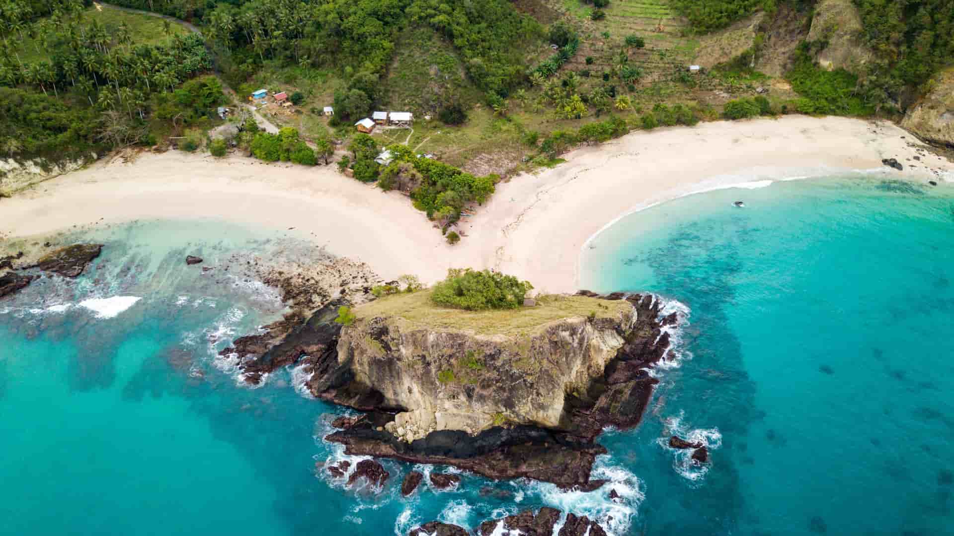 An aerial view of a picturesque twin-bay beach in Kokas, Indonesia, with clear turquoise water, a rocky outcrop separating two stretches of white sand, and lush green hills.