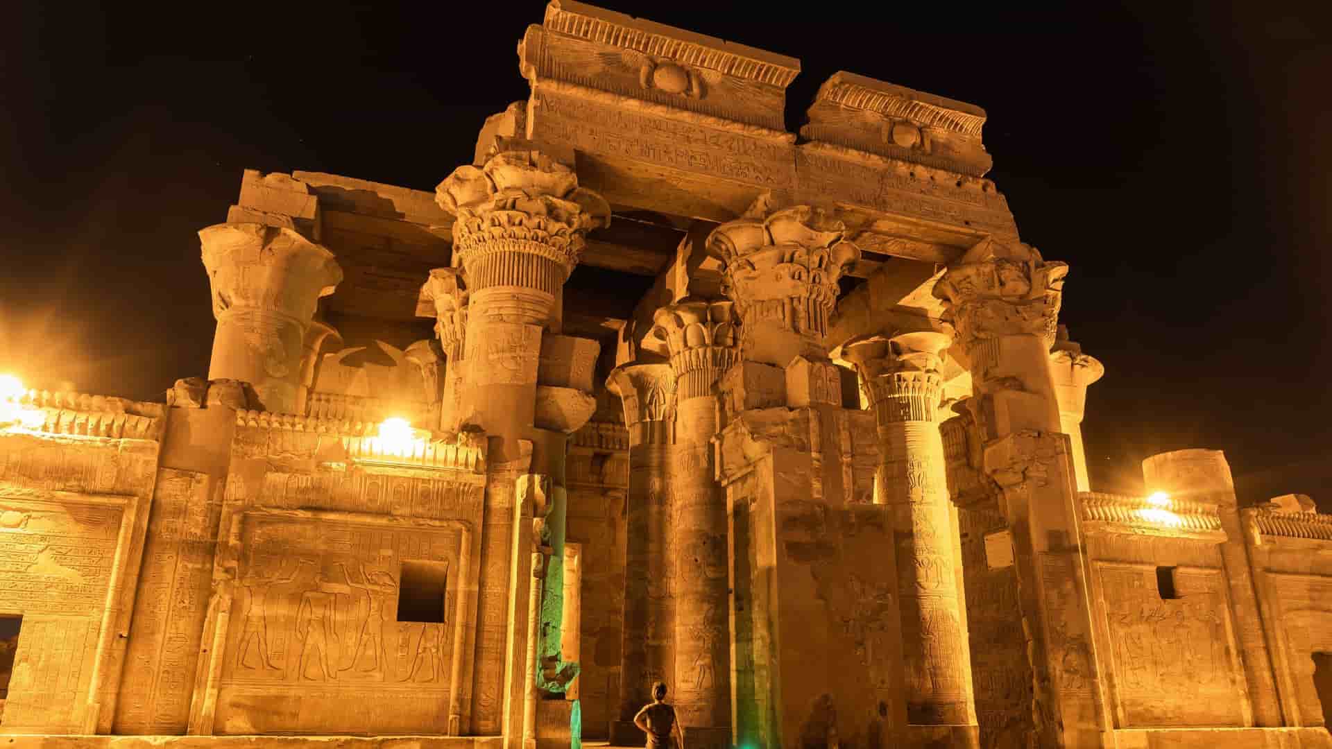 The magnificent Temple of Kom Ombo, an ancient Egyptian temple illuminated at night, with towering columns, intricate hieroglyphs, and a person standing in awe.