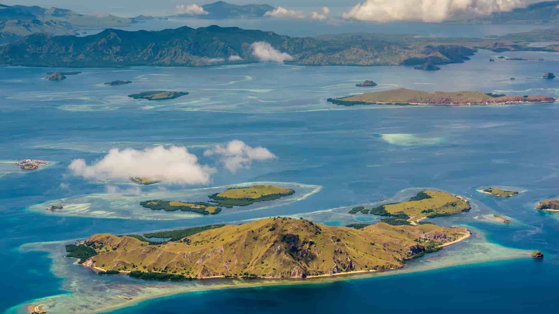 A stunning high-angle aerial view of Komodo Island and Batu Moncho in Indonesia, featuring lush green islands surrounded by vibrant turquoise waters and visible coral reefs under a bright sky.